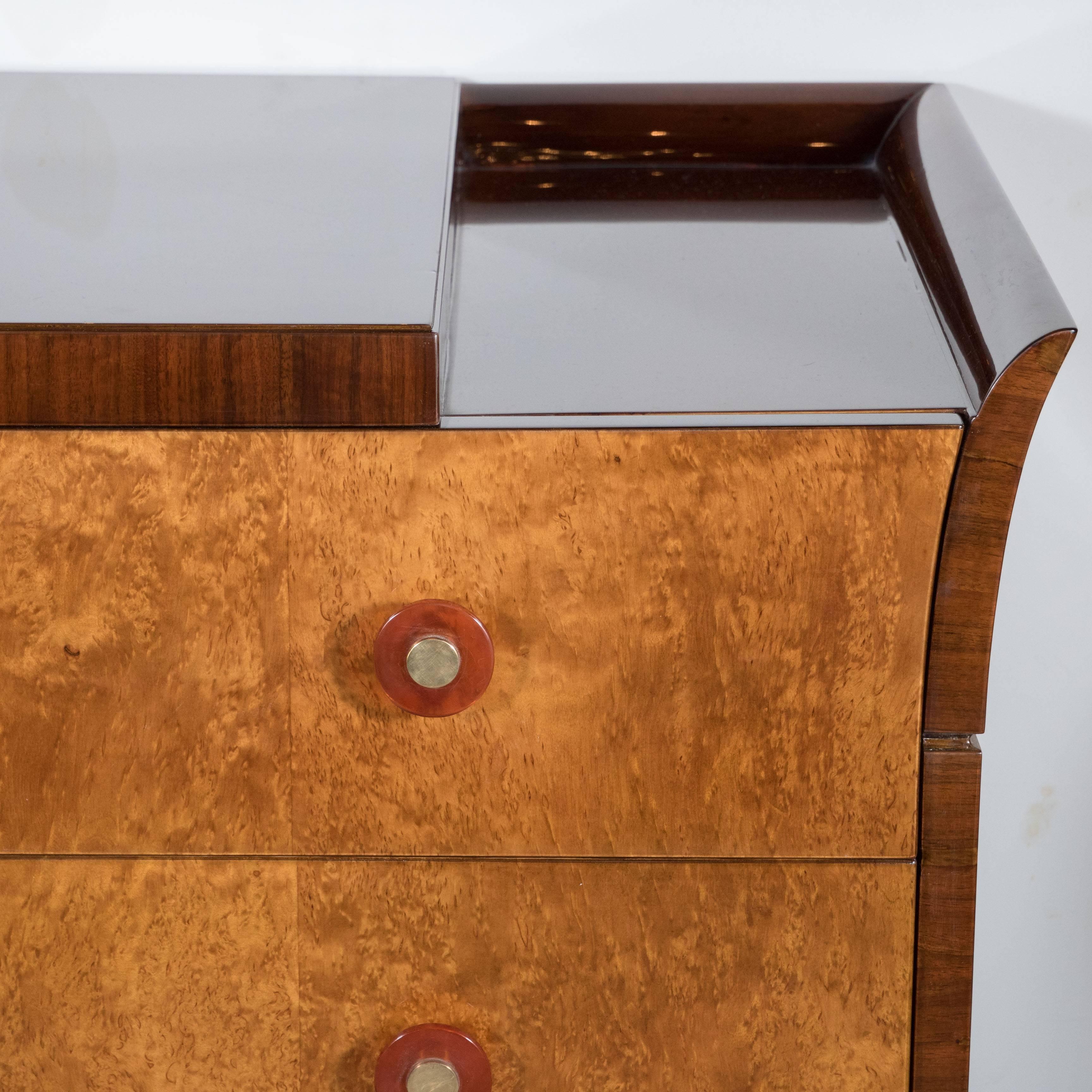 An Art Deco Skyscraper style chest featuring book-matched burled carpathian elm drawer fronts with amber bakelite and brass pulls, the top of the chest is book-matched mahogany with a raised platform positioned centrally to the piece surrounded by a