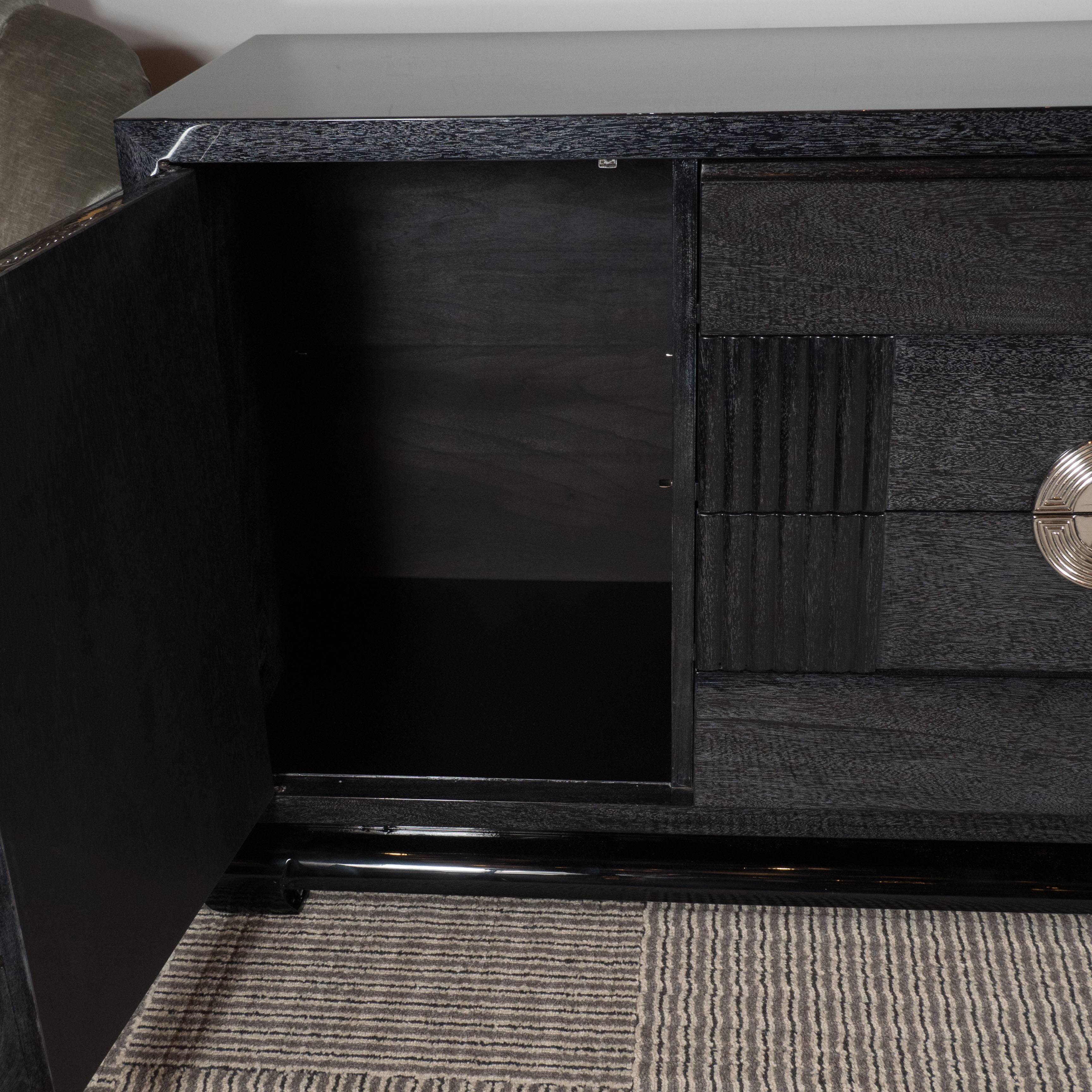 A Mid-Century modernist silver-cerused oak sideboard or cabinet with nickeled pulls, in the manner of Tommi Parzinger. A stepped base supports a pair of push-open side doors and three central drawers. Bevelled panels frame the side portions while