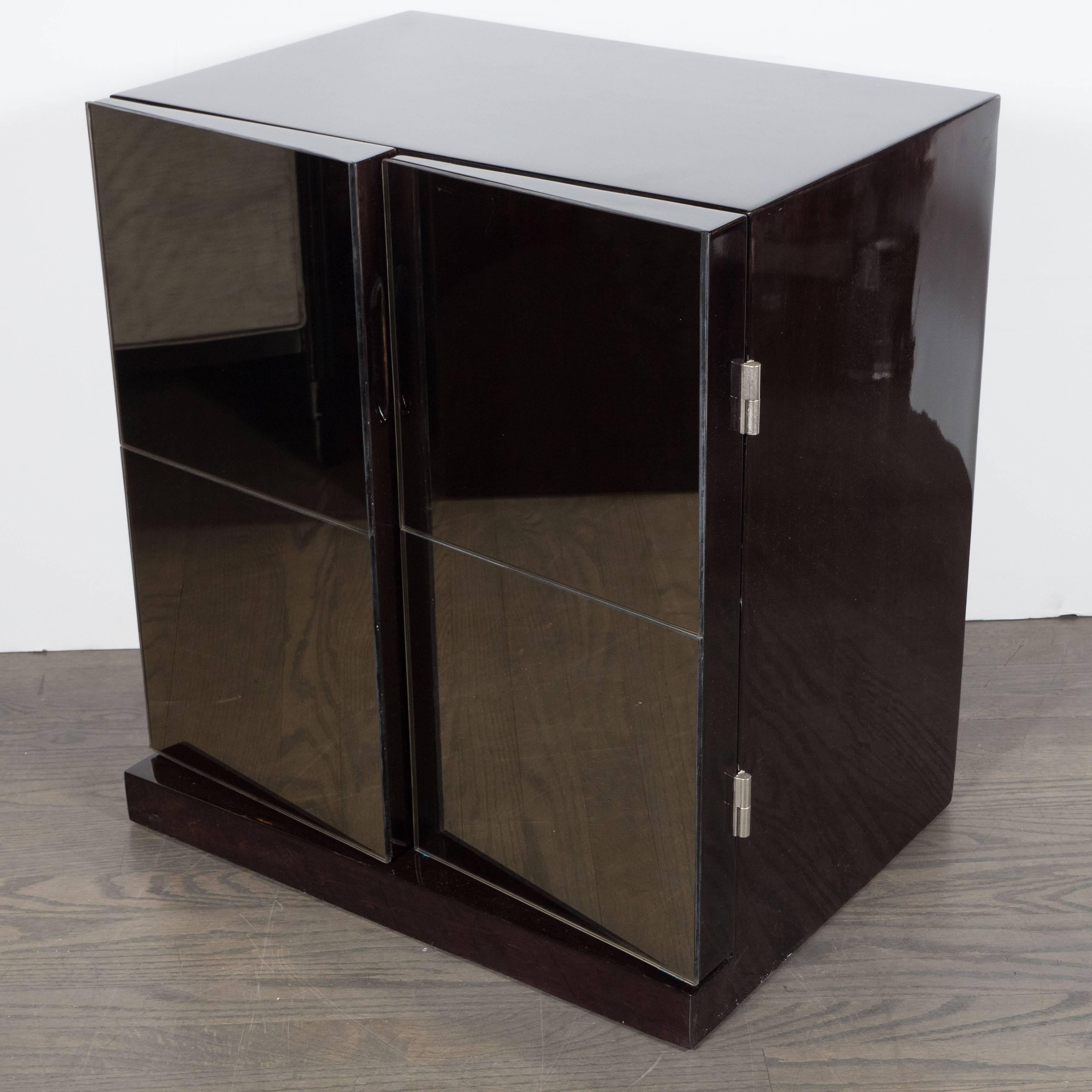 A pair of Mid-Century Modernist nightstands featuring angled Cubist smoked mirror fronts. Each piece of this mirrored pair features swing open doors revealing two levels divided by a single shelf. Finished in ebonized walnut, this pair could also be