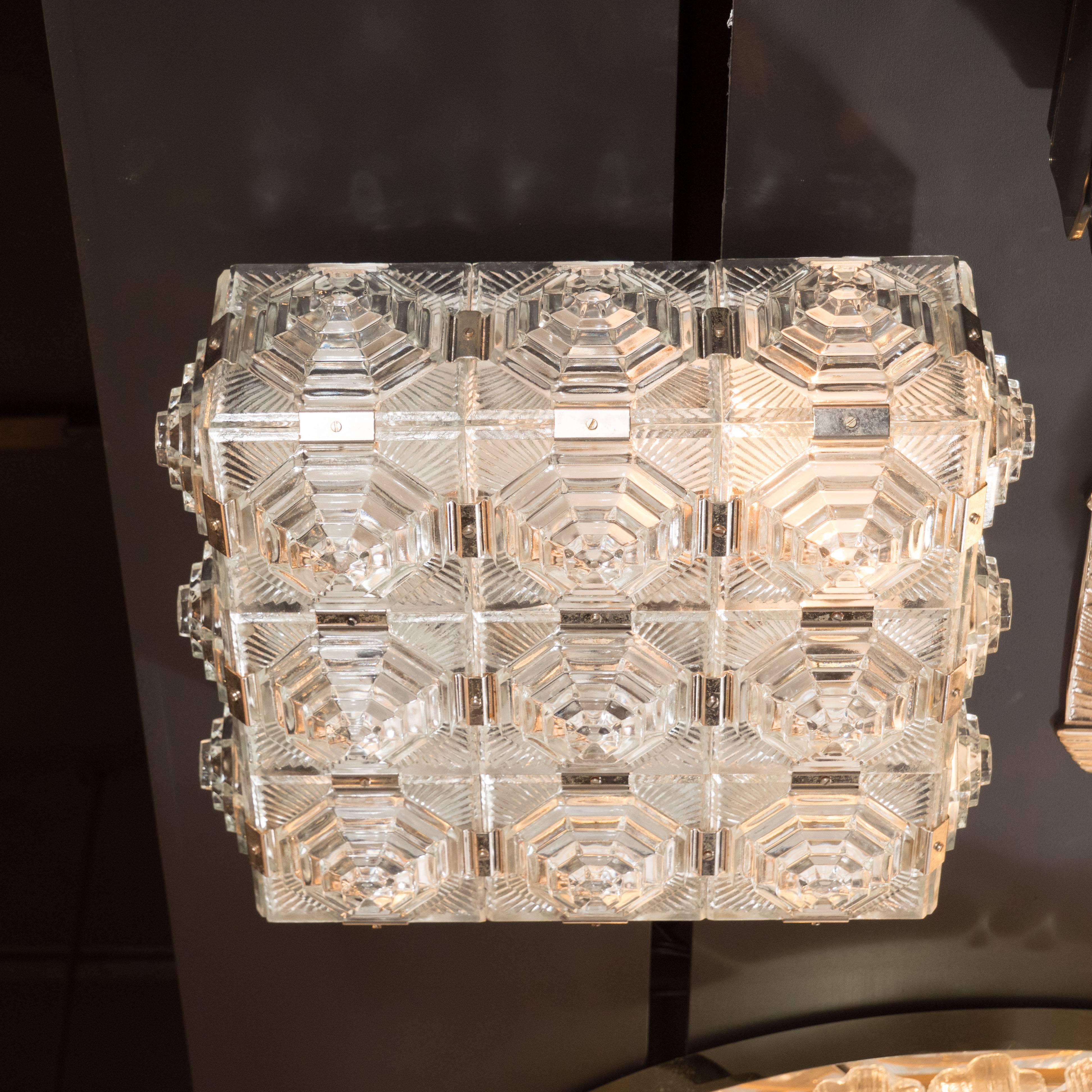 This stunning Mid-Century Modernist flush mount chandelier features an intricate pressed glass stylized geometric design and polished chrome fittings. There are nine glass panels adorning the bottom of the fixture, each inscribed with elaborate