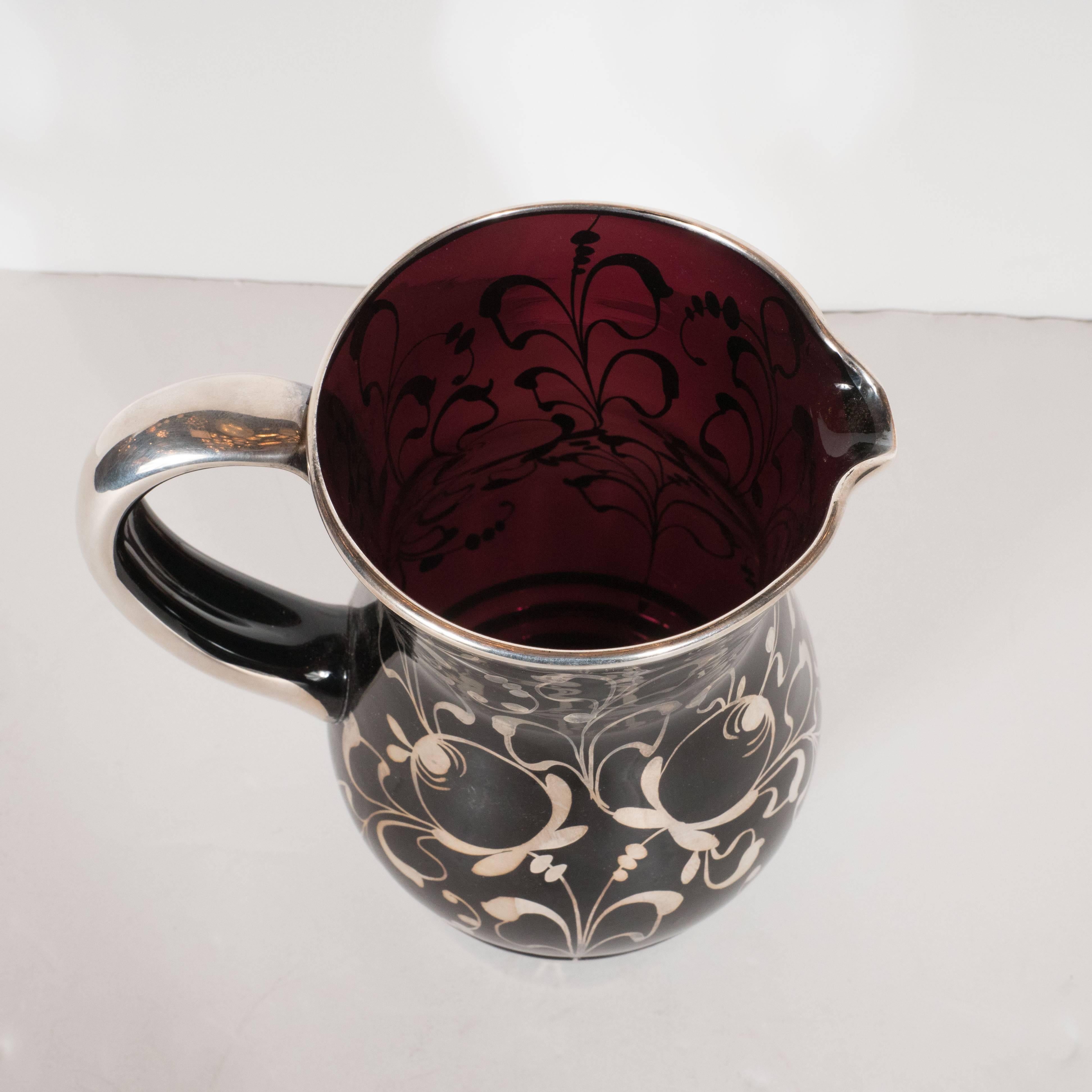 This handblown black glass pitcher features a stylized floral and fauna detailing and a ringed base overlaid in sterling silver. Additionally, the handle and rim have been overlaid in silver, making this a most elegant water, iced tea or seasonal