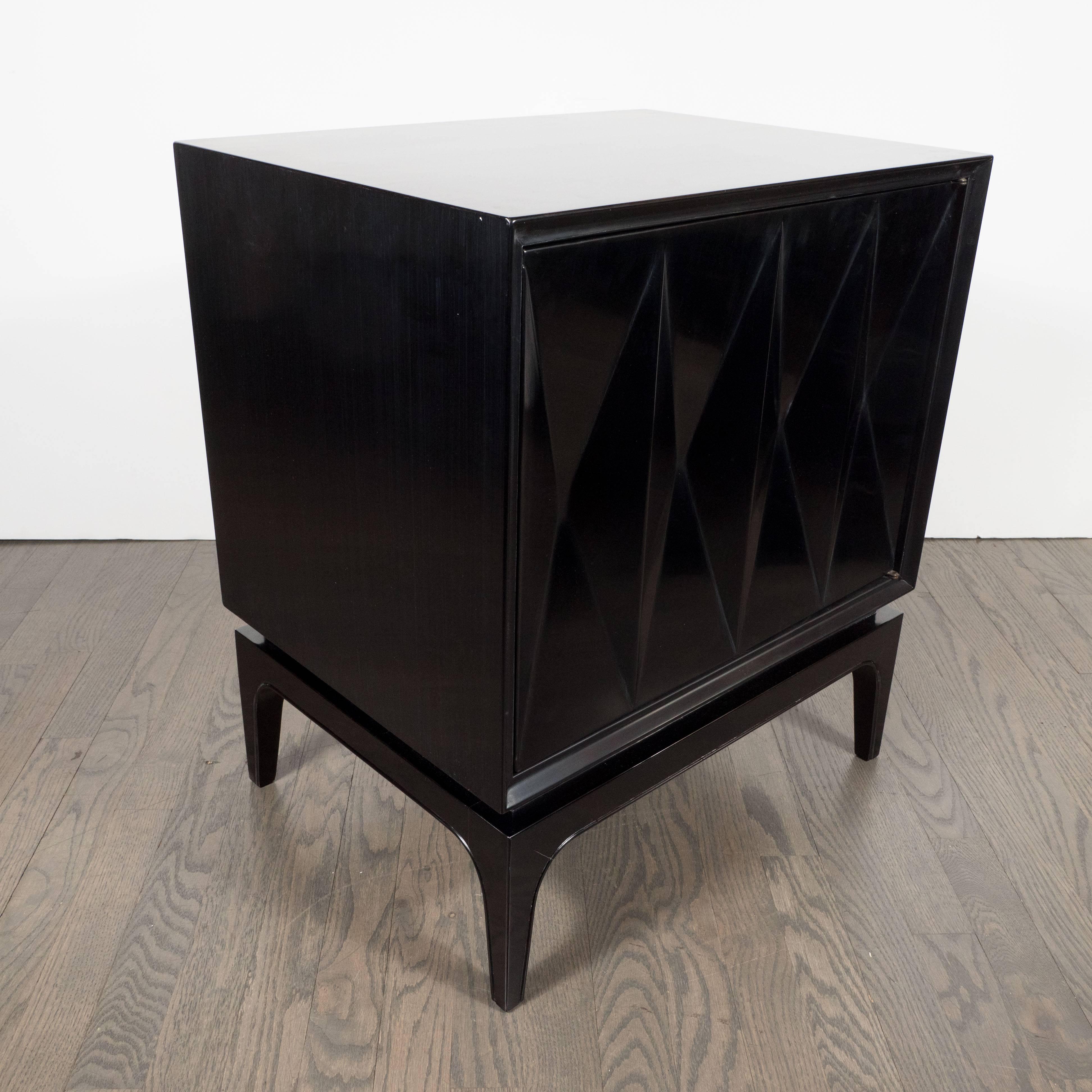 This sophisticated pair of ebonized walnut end tables or nightstands feature four rectangular panels with a concave cut triple diamond front panel, brass fittings, tapered conical legs, and an interior cabinet with one glass shelf. This pair