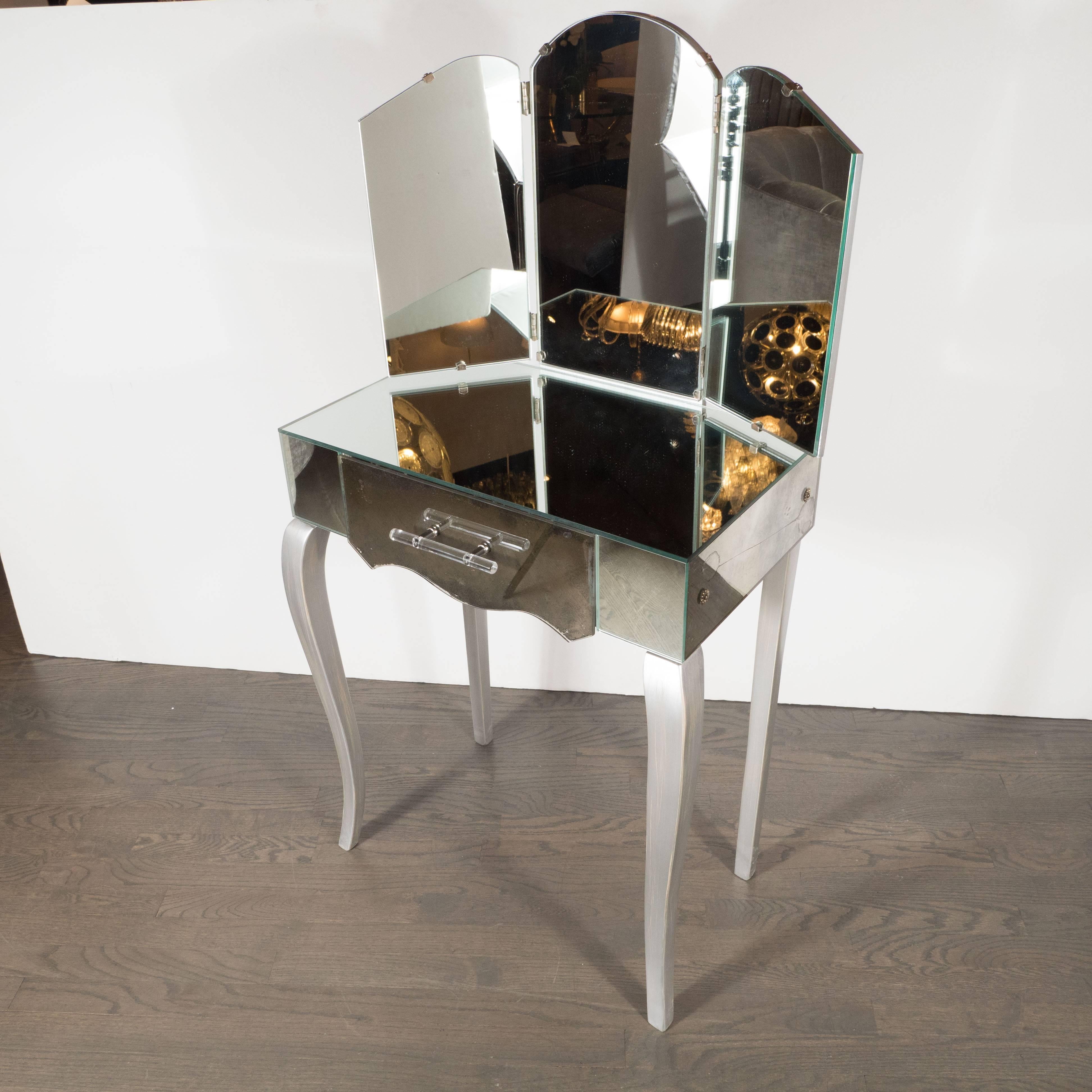 This posh 1940s Hollywood vanity features smoked/antique mirrored glass, floral rivets, and a Lucite and chrome pull. The front legs are cabriolet style, while the hind legs are straight and tapered. The entire back of the piece, the inside of the