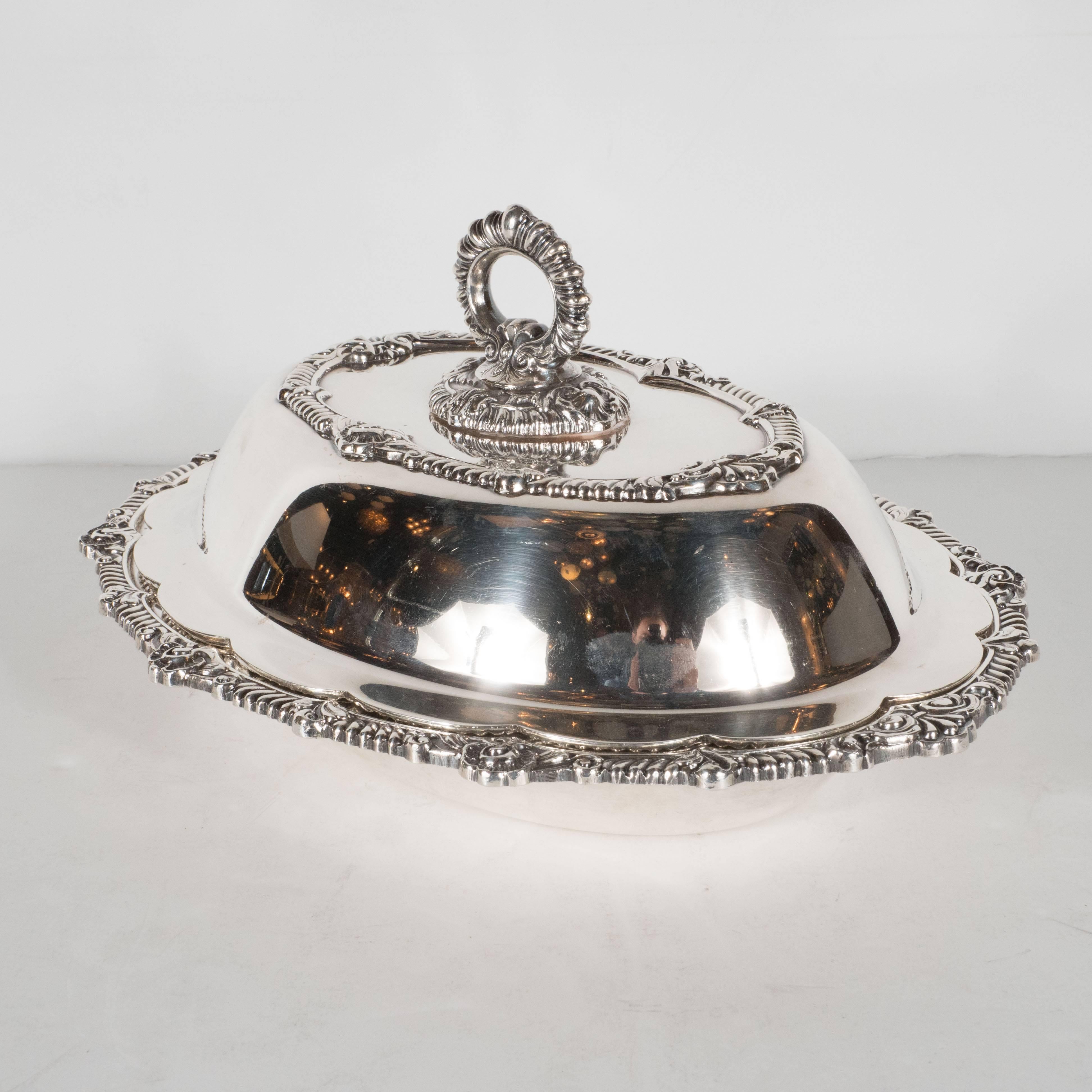 This stately sterling silver plated tureen features a reeded handle, scalloped edges with Baroque details, including fleur de lys throughout. The lid features a reeded ovular handle and a raised based with intricate Baroque designs. The tureen comes