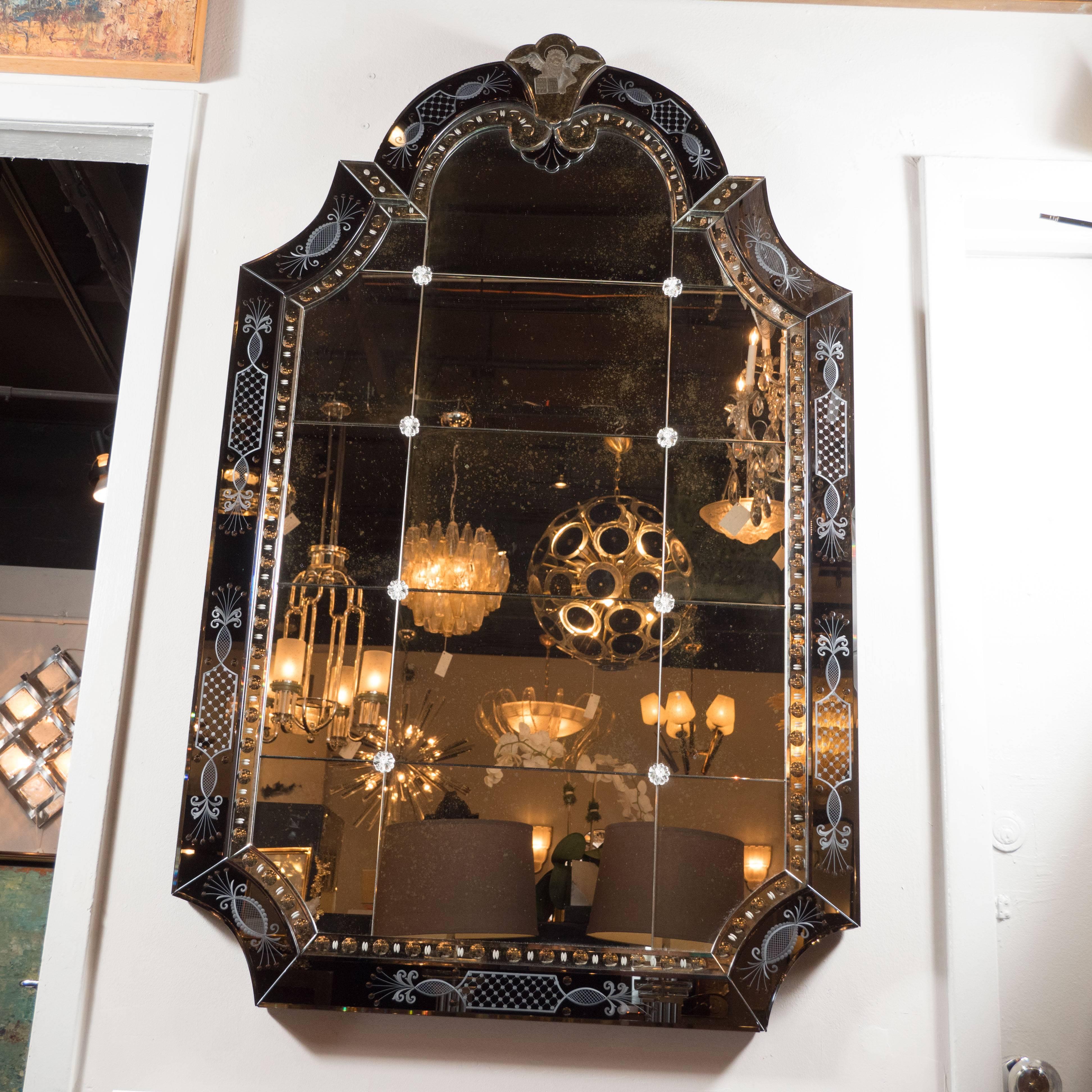 This elegant Hollywood Regency/Art Deco Mirror features a mosaic of twelve rectangular panels and one arched top panel fastened together with glass floral rivets inside of a scalloped, beveled, and smoked mirror frame with reverse etched Baroque
