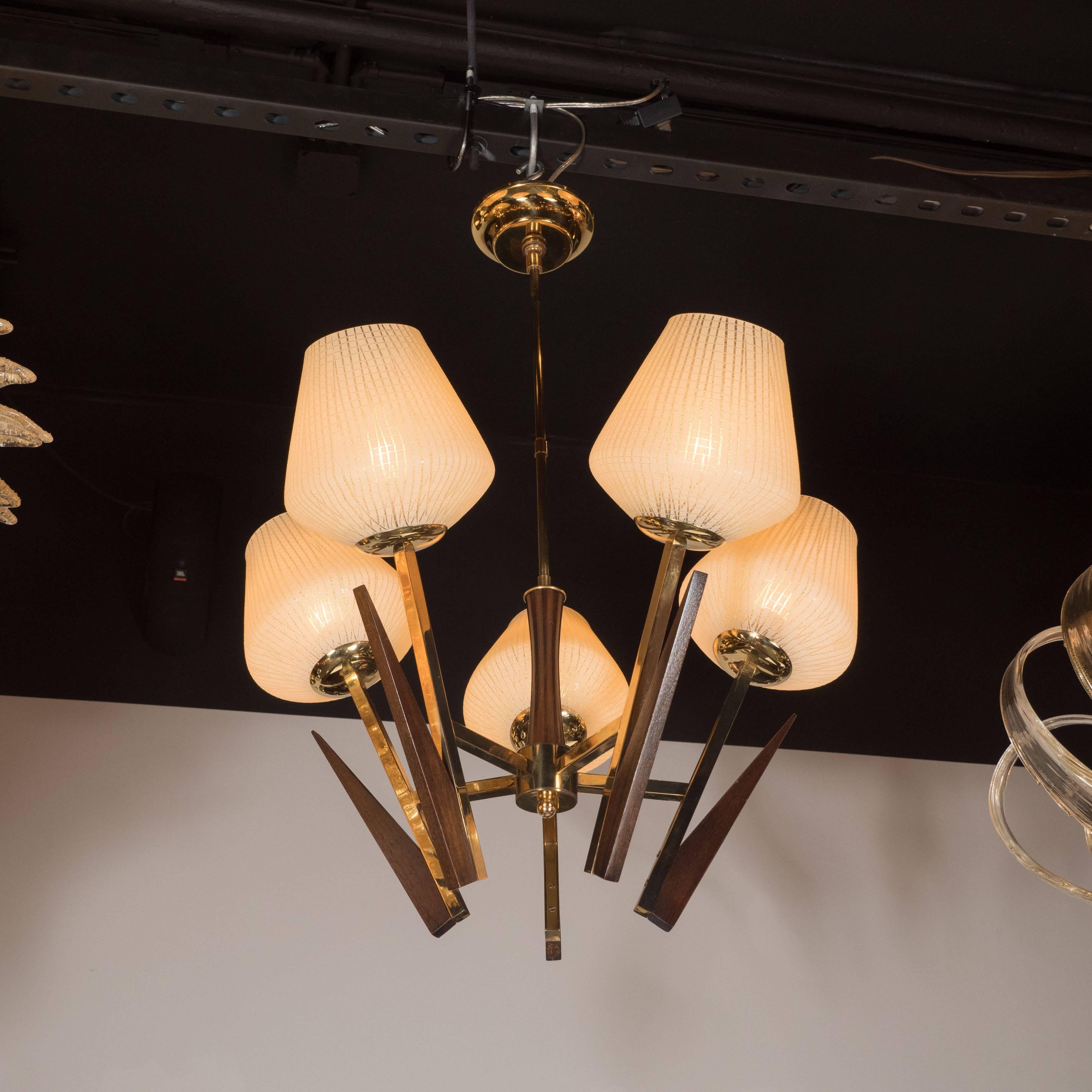 This sophisticated chandelier features five frosted ecru glass globes with clear cross hatching in organic geometric patterns and a billowing form that tapers towards the top. The angular and geometric forms of the arms in lustrous brass with