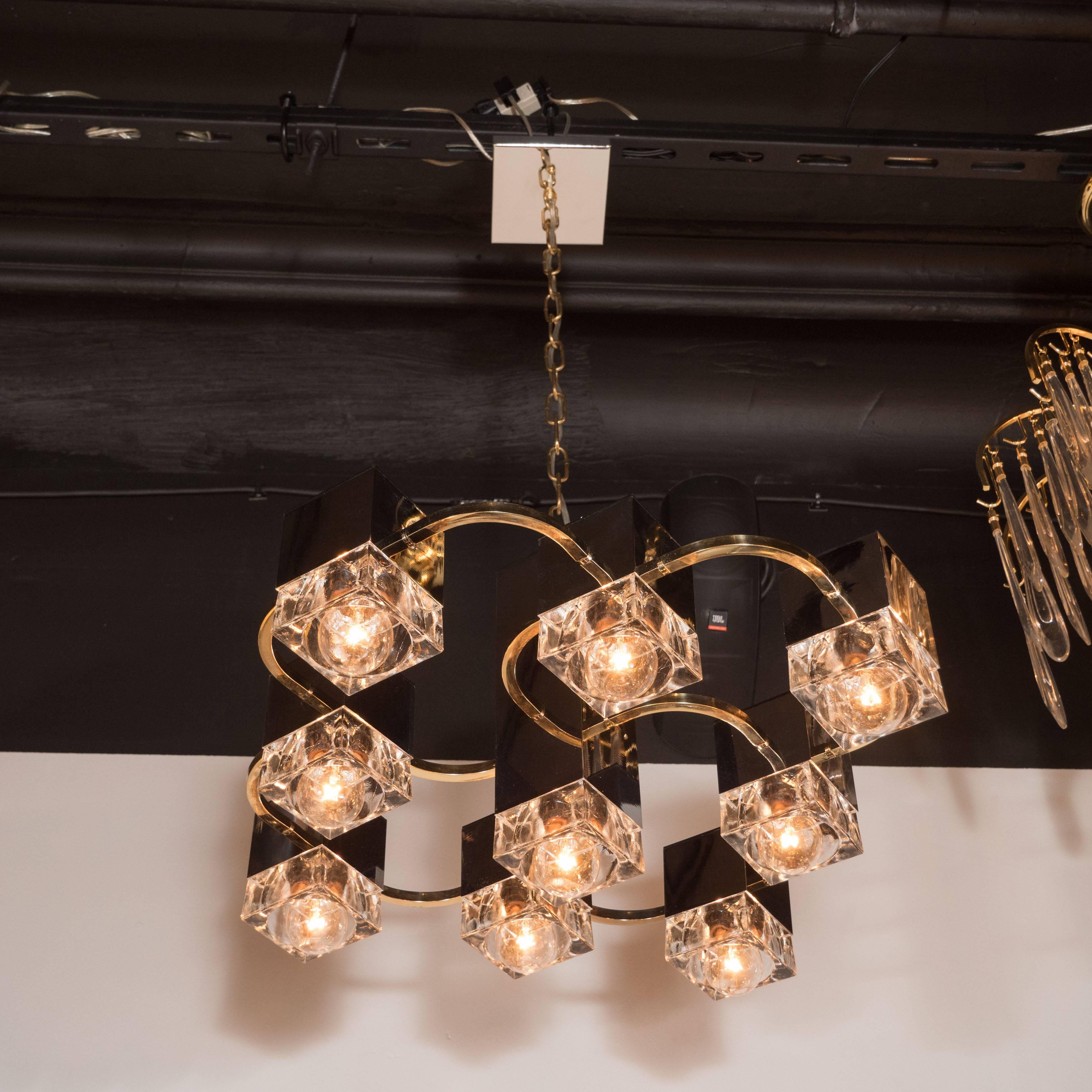 This sophisticated Scolari chandelier features a central rectangular pillar in chrome with eight chrome cube forms, holding waterglass fittings that attach to curved brass arms. This fixture embodies sophisticated Mid-Century Modern design at its