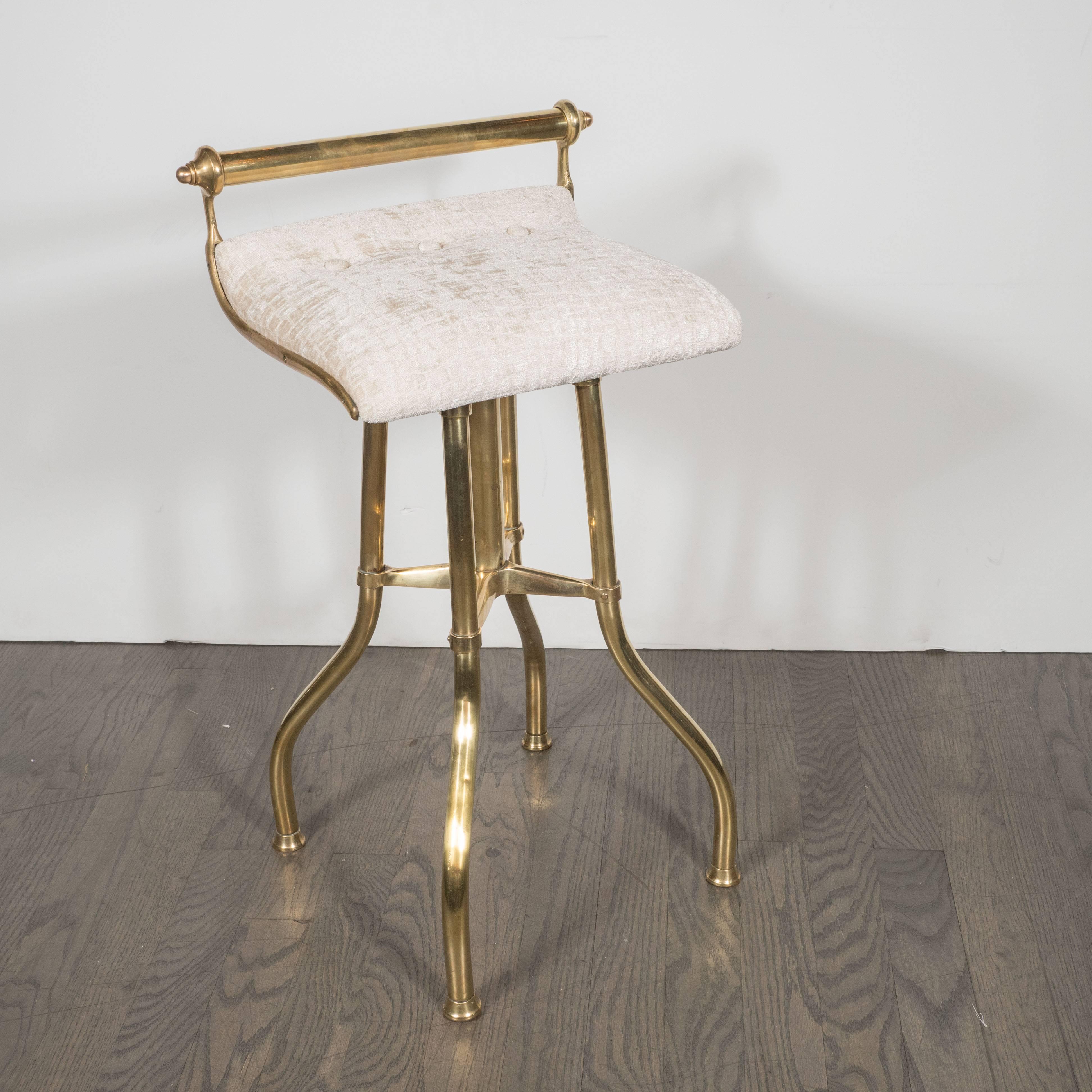 This glamorous Mid-Century Modern vanity stool features a cylindrical back support with circular ziggurat ends in lustrous brass and four legs of the same material that descend from the seat straight before flaring out at the base. Brass cuffs