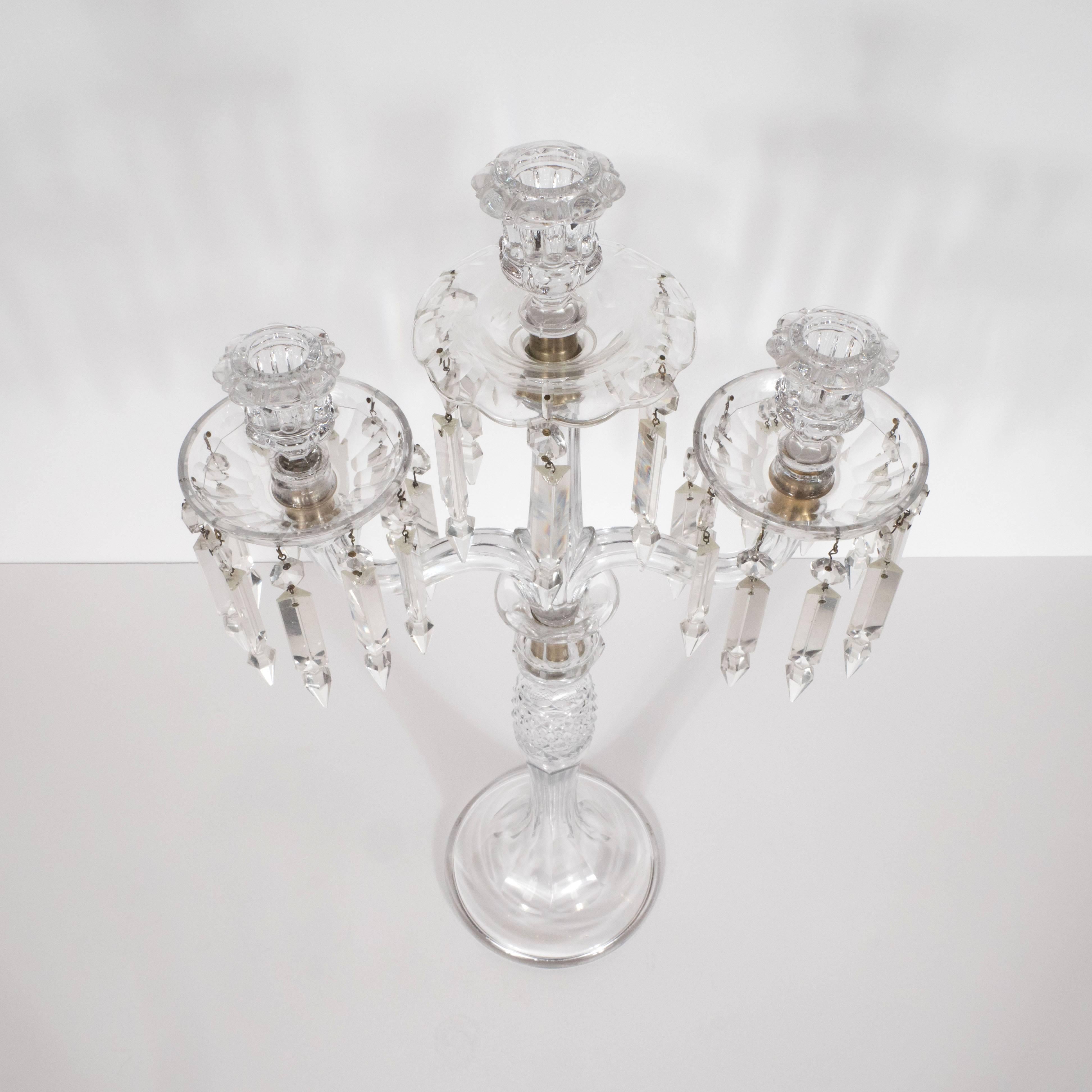 This exquisite pair of crystal girandoles features a central stem with two scroll form arms branching off to support convex canopies to catch melting wax and stylized floral candleholders. The central stem offers a circular base and a reeded pattern