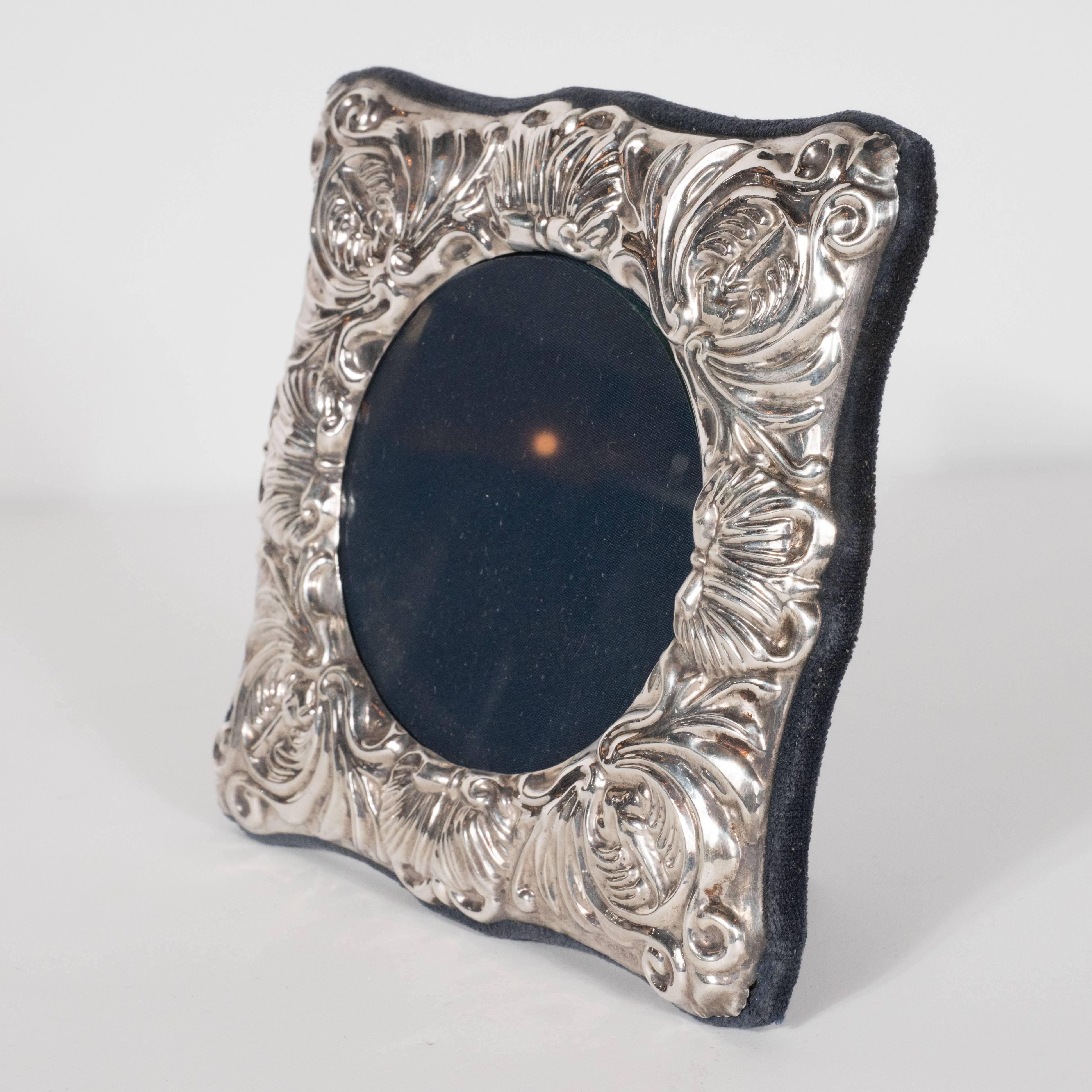 This charming picture frame features a circular glass sheet in the centre, surrounded on all sides by Baroque detailing in sterling silver repoussé. The back is composed of midnight blue velvet. This is a fine example of English sterling, not to