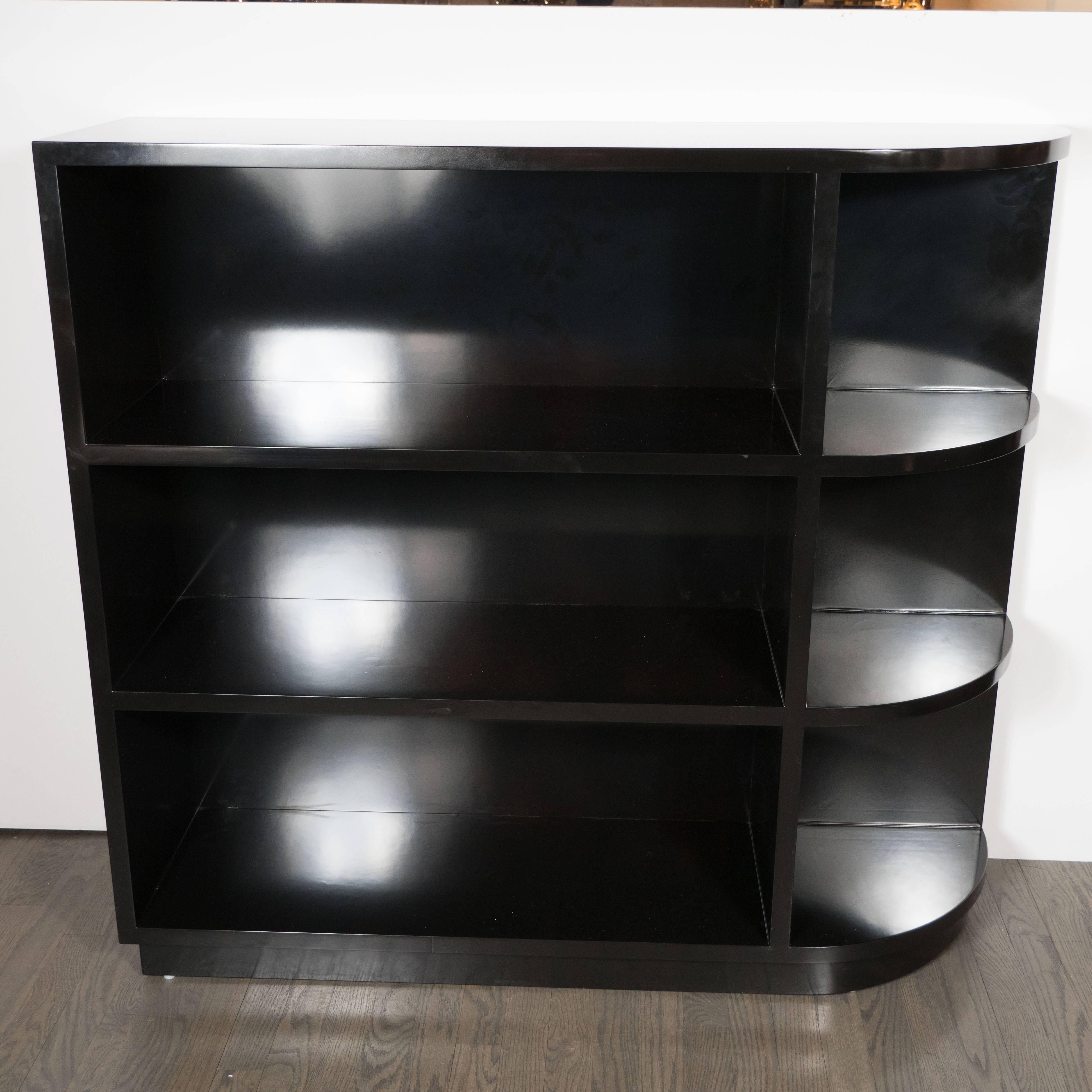 A striking American Art Deco black lacquered chest level bookcase with one straight end and another streamlined end, complete with two book shelves spanning the entirety of the piece and smaller semi-circular shelves ideal for decorative objects. It