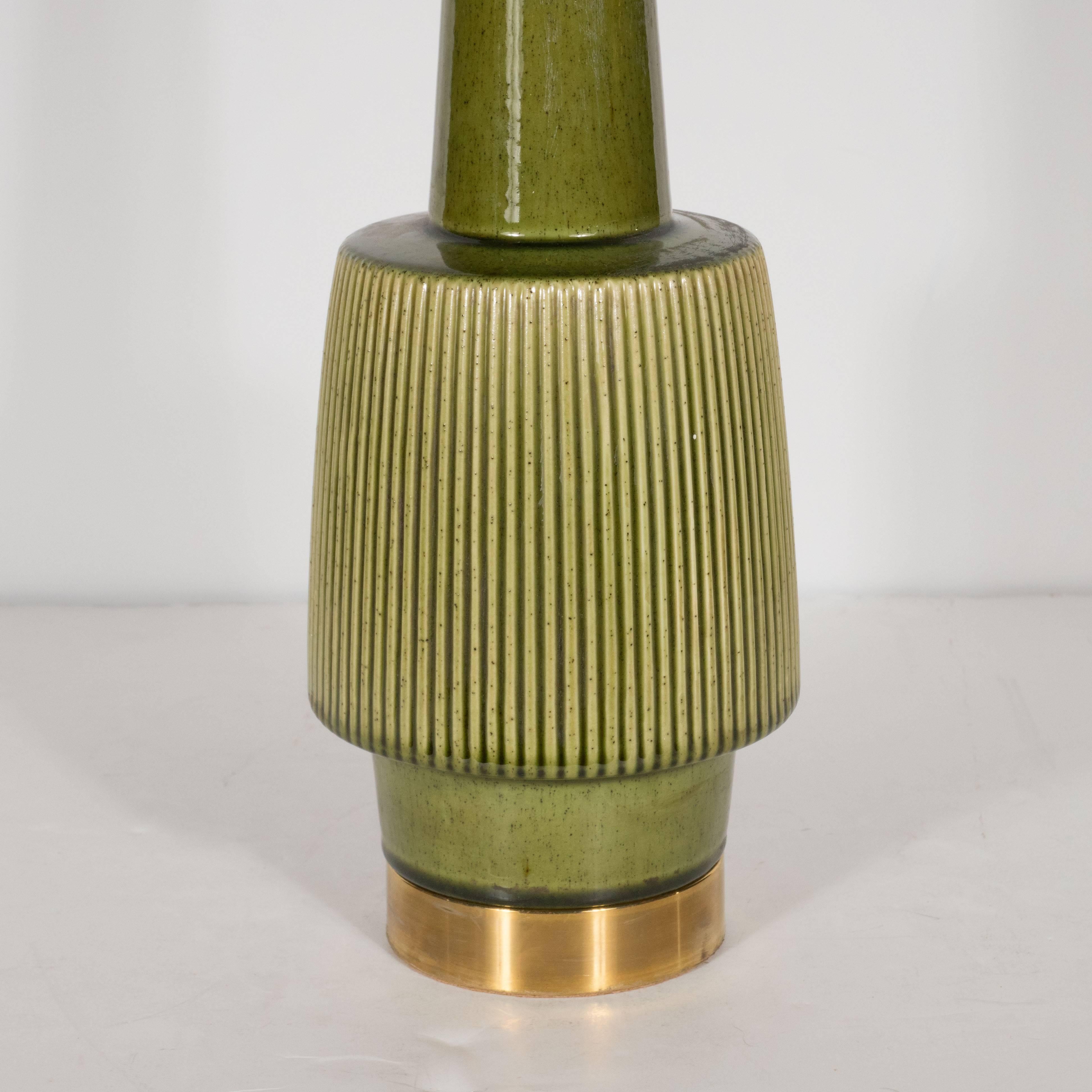 This pair of Mid-Century Modern table lamps were handcrafted in ceramic and glazed in a finely speckled green olive hue. The lamp rests on a brass base which ascends into a cylindrical form on which a larger striated form of the same shape sits. A