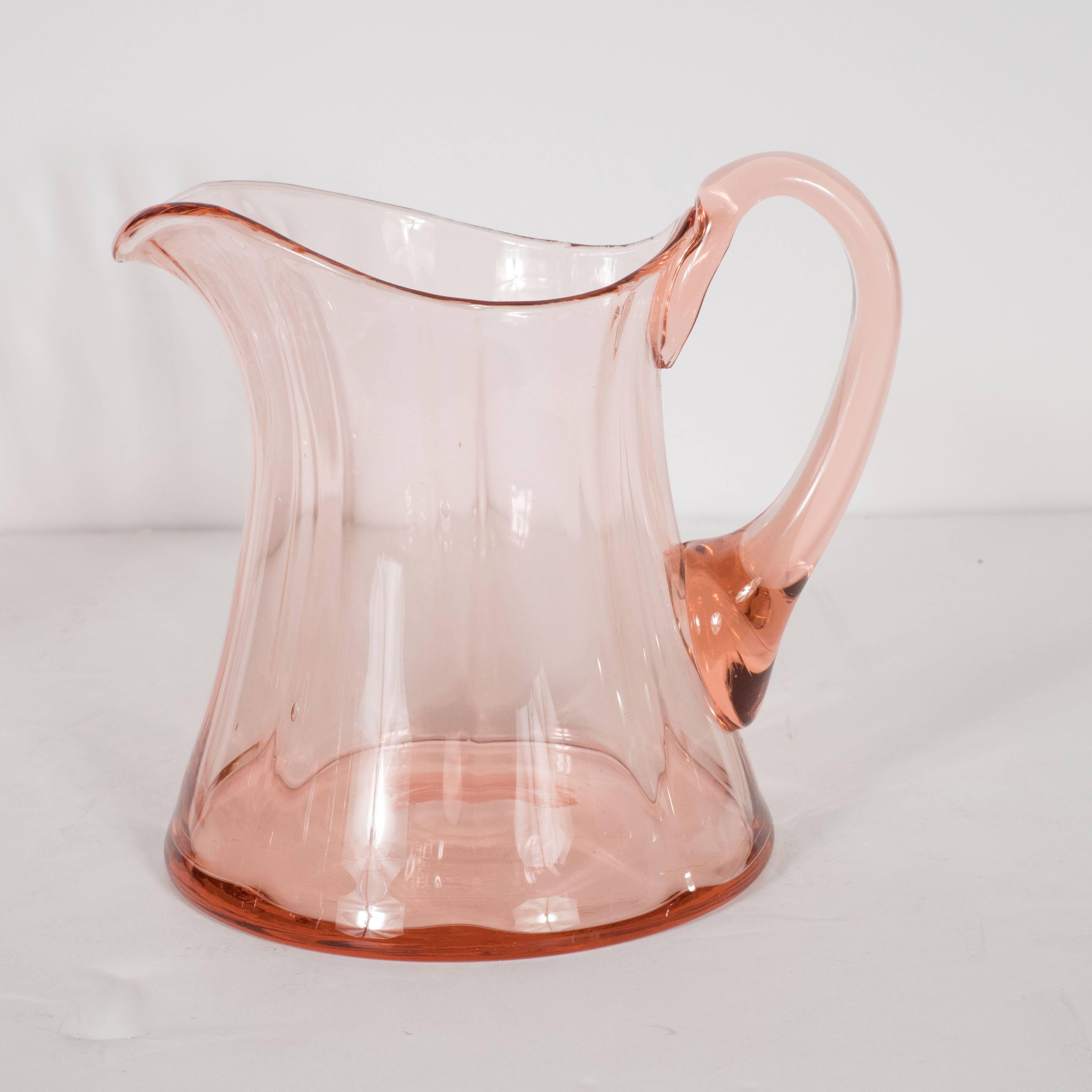 The pitcher offers a billowing channeled form with a gently curved front culminating in an undulating lip blown in a blush rose hue. This pitcher would be perfect for serving lemonade, ice tea, punch or any number of drinks. It represents a