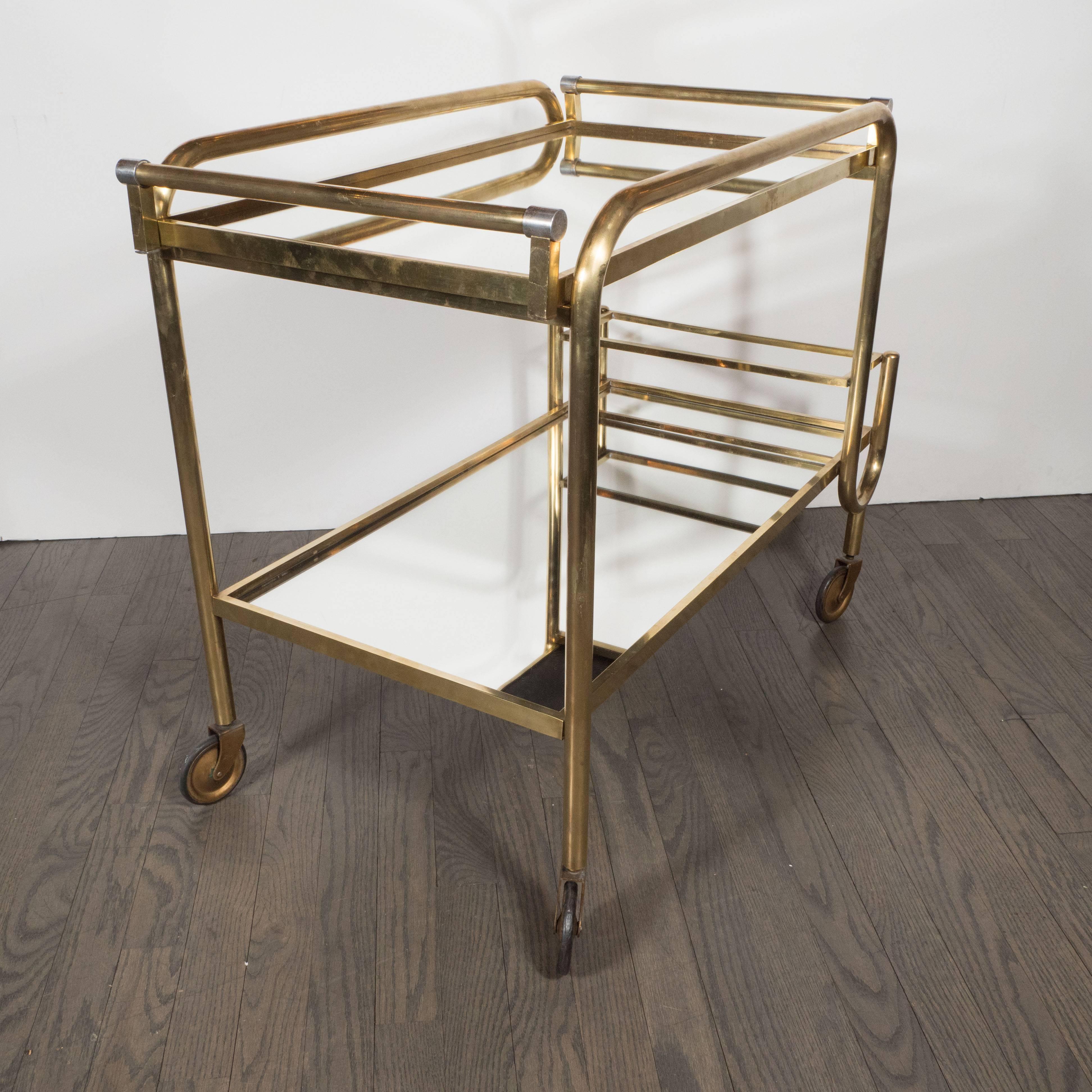 This stunning Mid-Century Modern brass and mirrored glass bar cart was realized in Italy, circa 1950. It features two tiers of mirrored glass- a tray on top, and a shelf on bottom- connected with an undulating tubular frame in brushed brass. At the