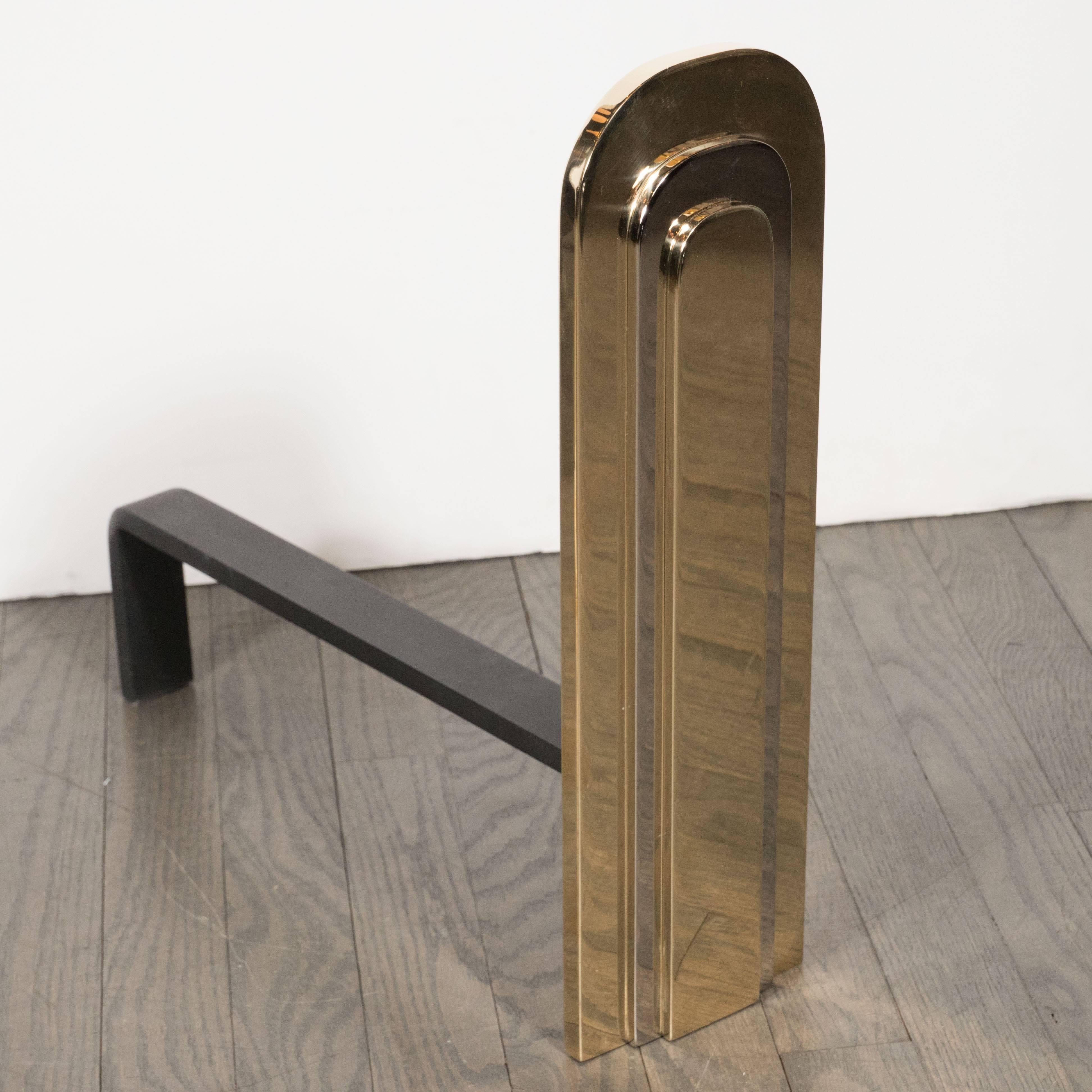 Handmade by artisans in New York state, this pair of modernist andirons embody the old world craftsmanship and design aesthetic of a bygone era. With their skyscraper style construction, consisting of alternating curvilinear tiers of polished brass