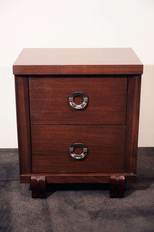 This stunning pair of Mid-Century Modern nightstands were realized in the United States, circa 1950. They feature two spacious drawers and stylized ring pulls with horizontal striations in nickel. Additionally, they offer scroll form embellishments