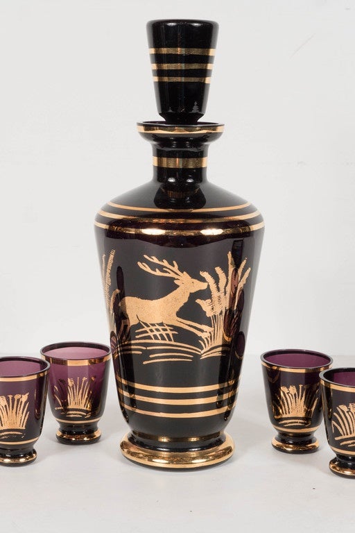 This stunning decanter set features a decanter and eight shot glasses in amethyst (almost black) glass with an Art Deco design in 24-karat yellow gold of a leaping gazelle with stylized foliage and fauna design with stylized concentric rings