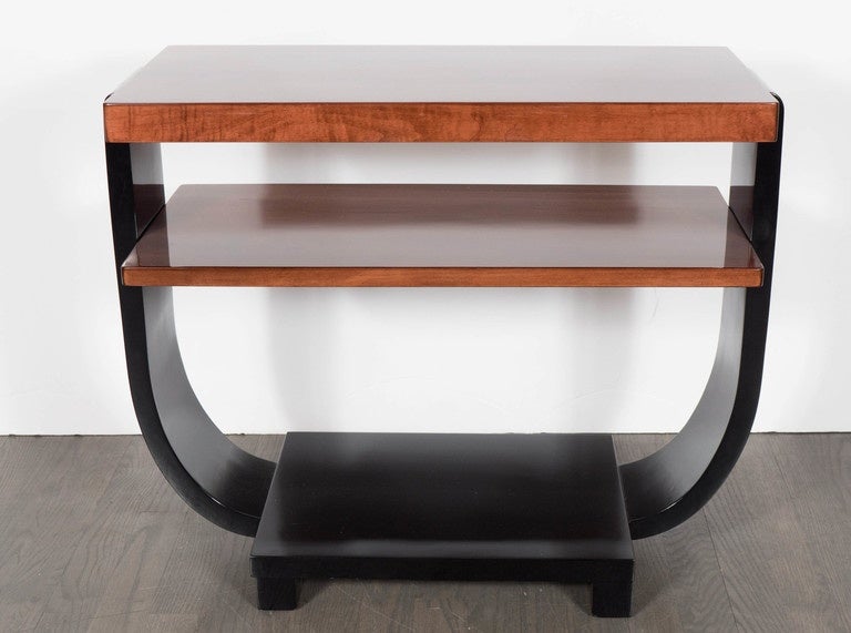 This beautiful table is a great example of the streamline Art Deco movement in America. It is made of bookmatched walnut and black lacquer and has two tiers with concave bentwood supports and lower shelf. It has been completely restored to mint