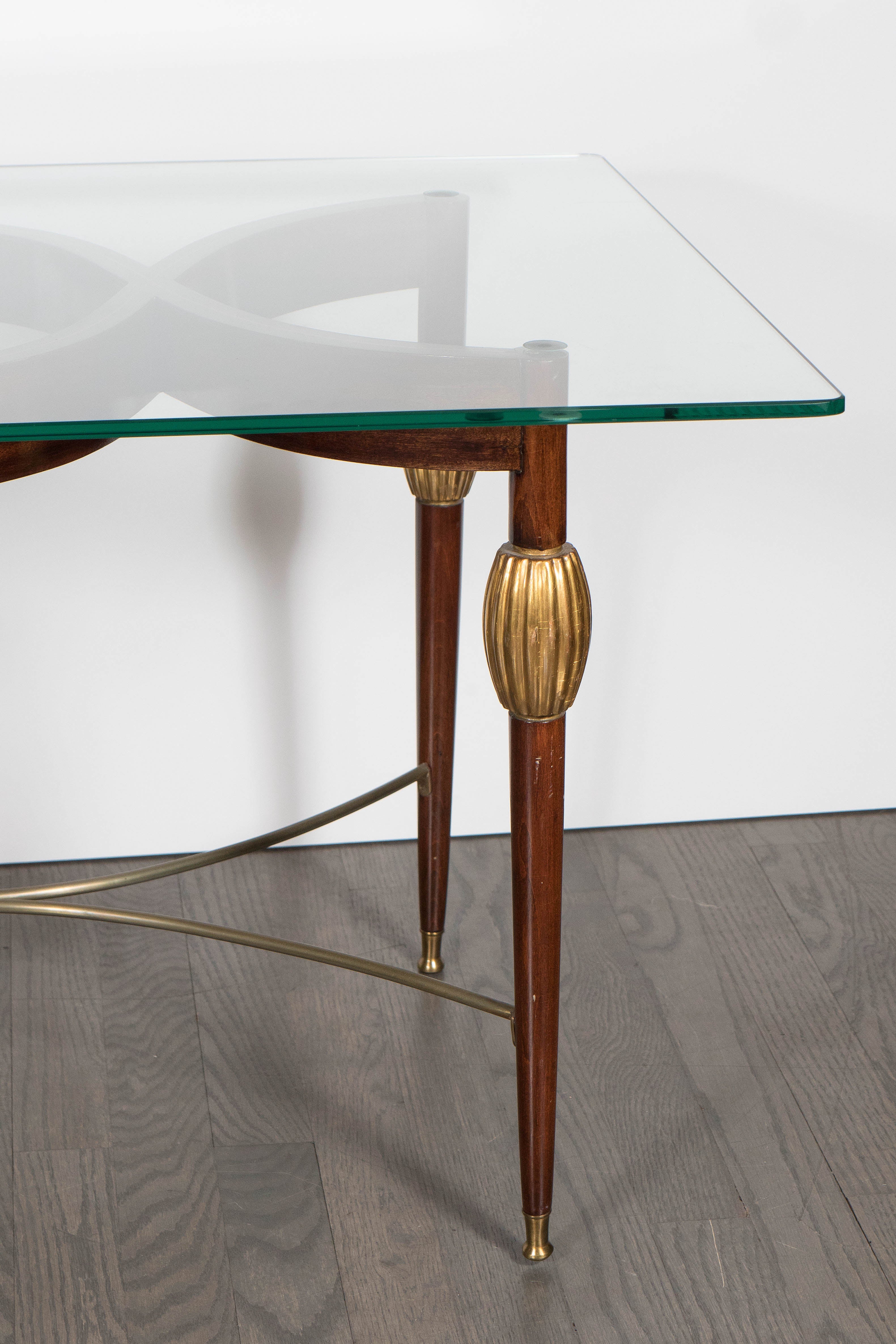 A stylish Mid-Century Modern Italian cocktail table in the style of Gio Ponti in mahogany and holly inlay with 24-carat gold giltwood and brass support and sabots. The simple yet elegant transparent glass tabletop lays on the delicately curvy shaped