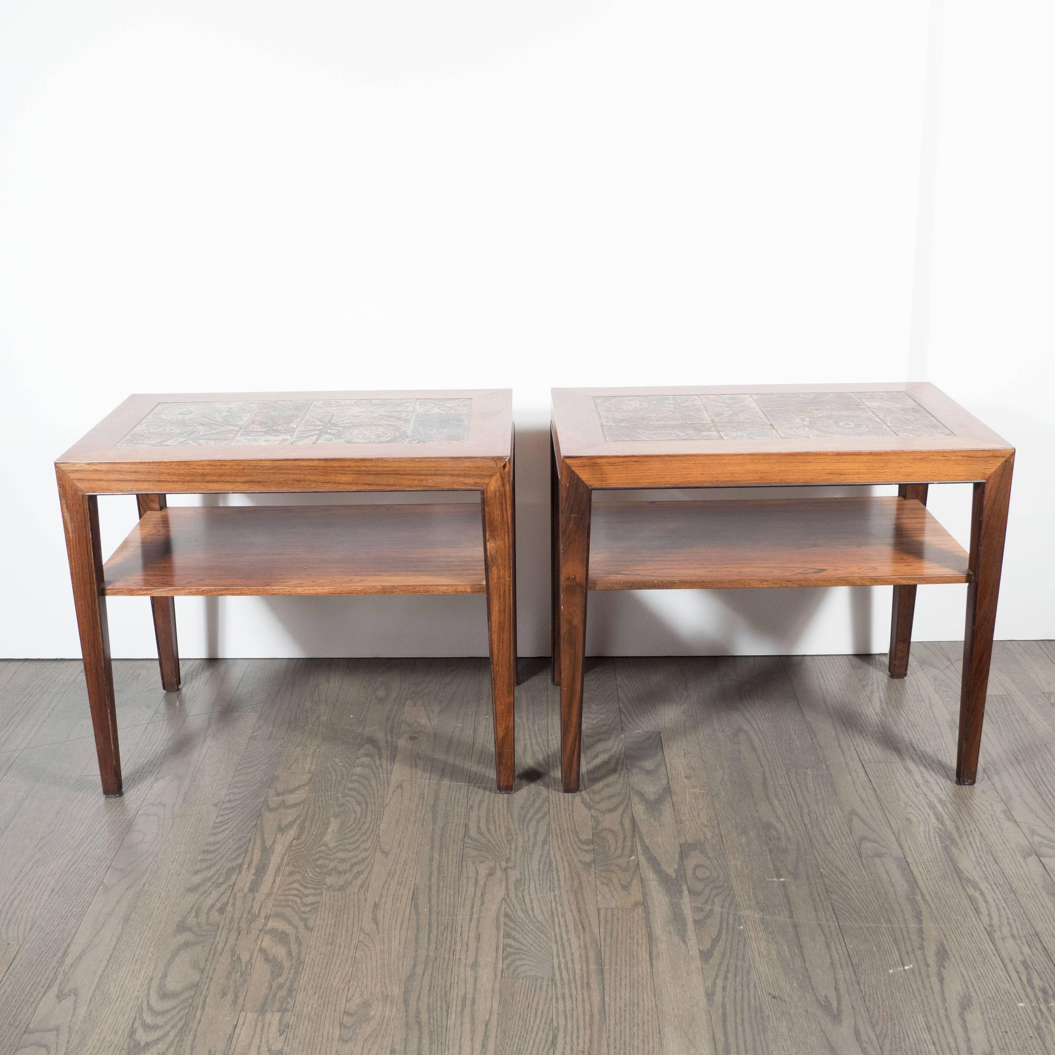 A pair of Mid-Century end tables in rosewood with inset hand-painted Royal Copenhagen tiles. Slightly tapered legs each hold a lower shelf. The tops of each display a series of hand-painted Royal Copenhagen tiles, featuring various earth-toned