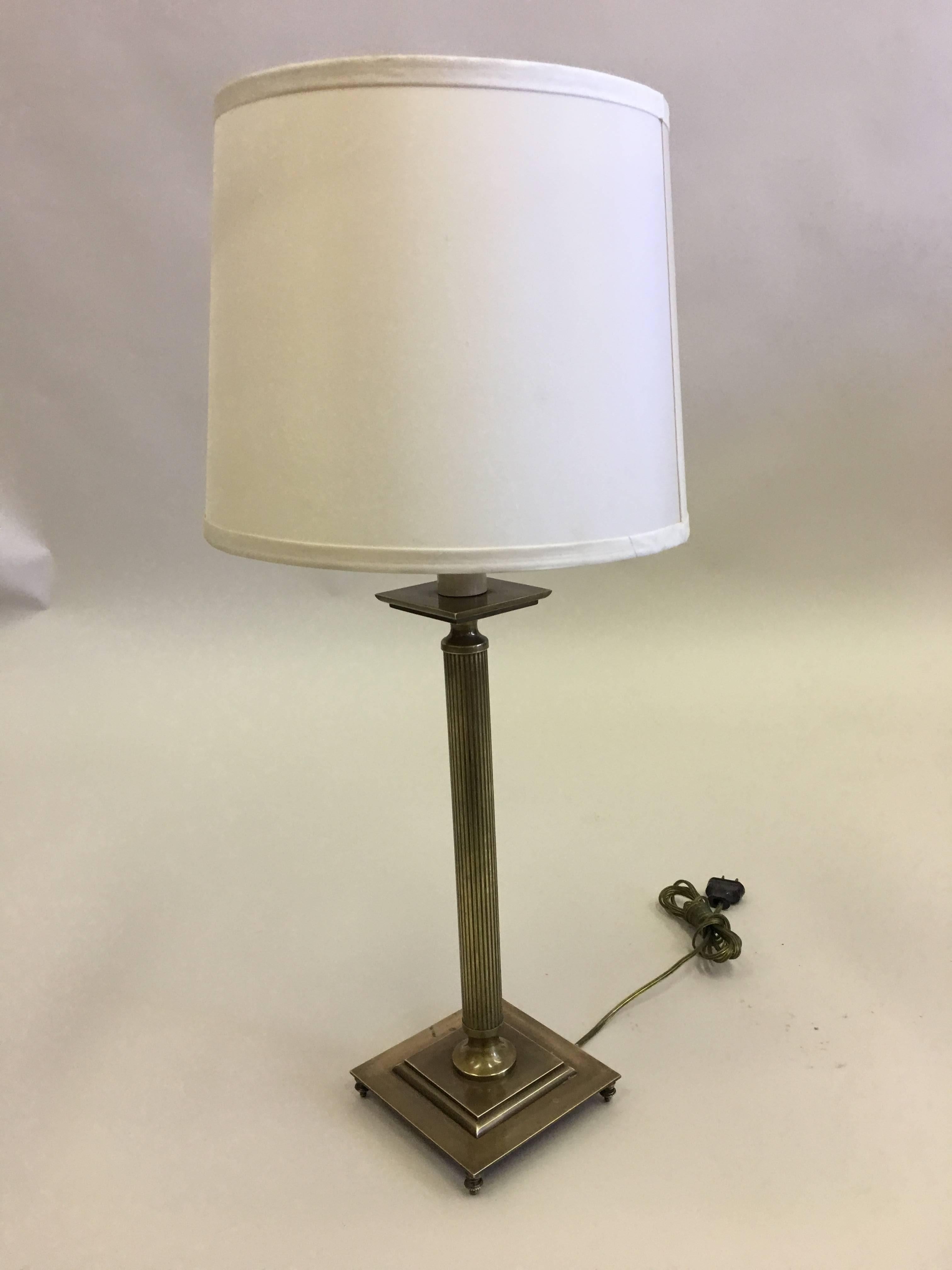 Elegant pair of French solid brass table lamps in the modern neoclassical tradition with fluted brass stems and raised brass square base resting on delicate brass finials. The pieces have a naturally aged antique brass patina. Shades are for