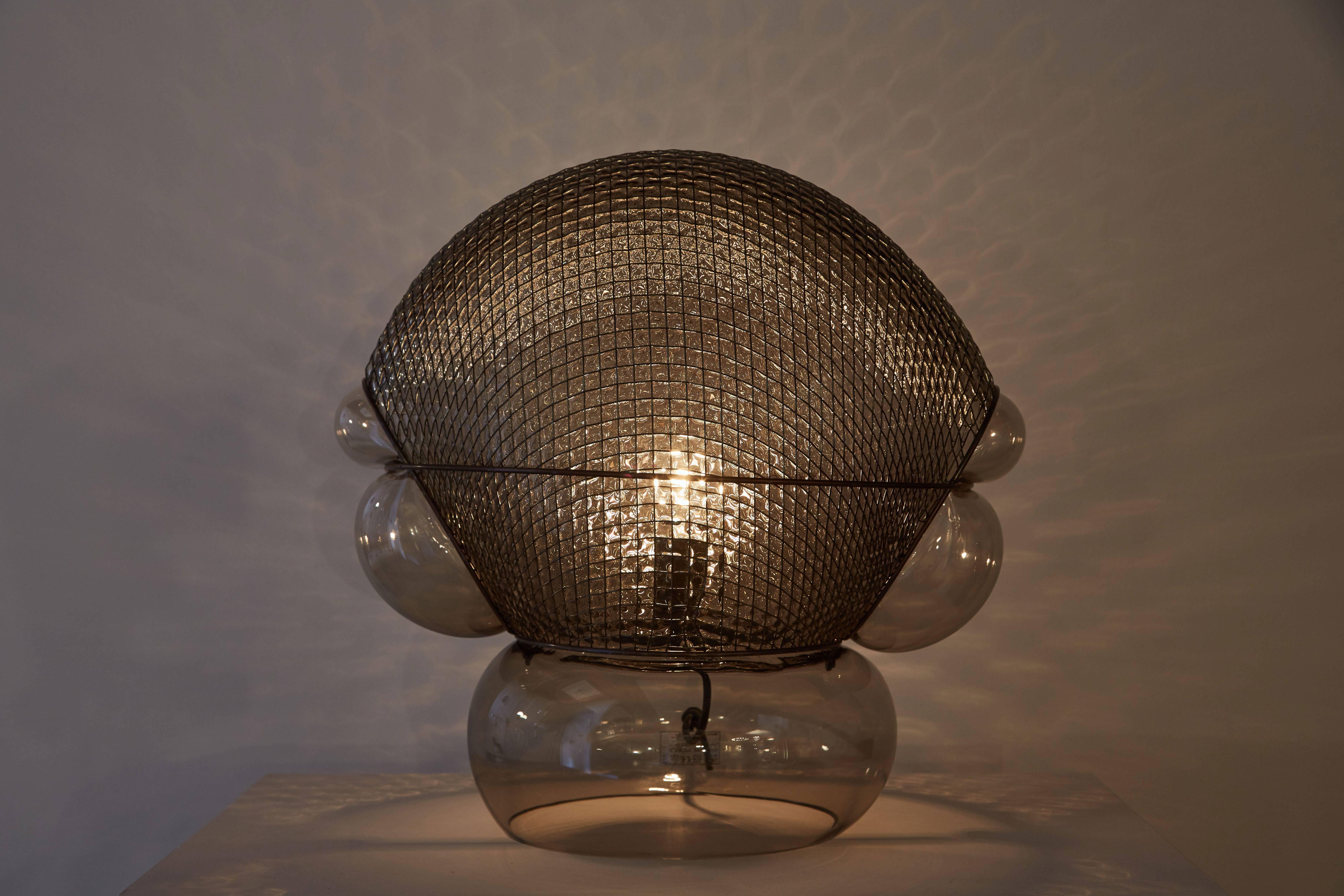 “Patroclo” table lamp by Gae Aulenti originally designed in 1975. Currently produced in Italy by Artemide. Comes with dimmer on the hand switch. Body made of smoke colored blown glass and steel mesh. Maintains original Artemide label. Takes one E27