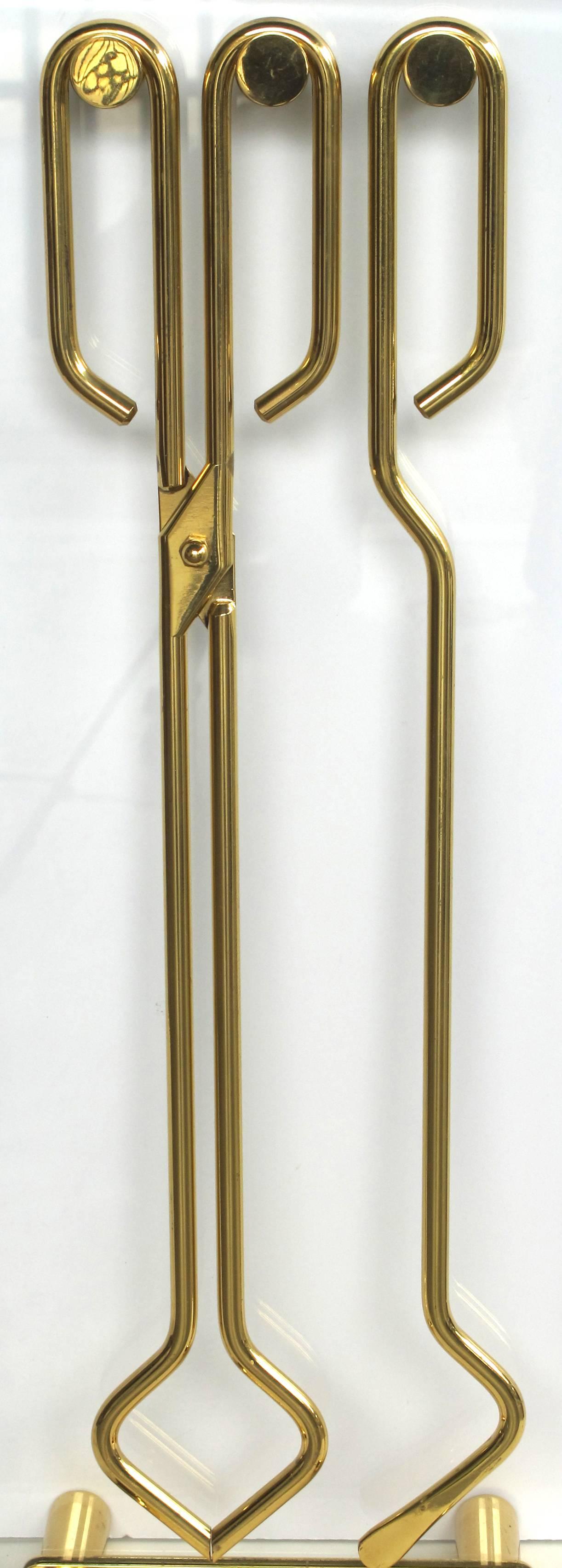 A stylish French 1960s gilt-metal and glass fire tool set in the manner of Jacques Adnet; the upright glass stand raised on cylindrical rods; with protruding knobs suspending two tools.