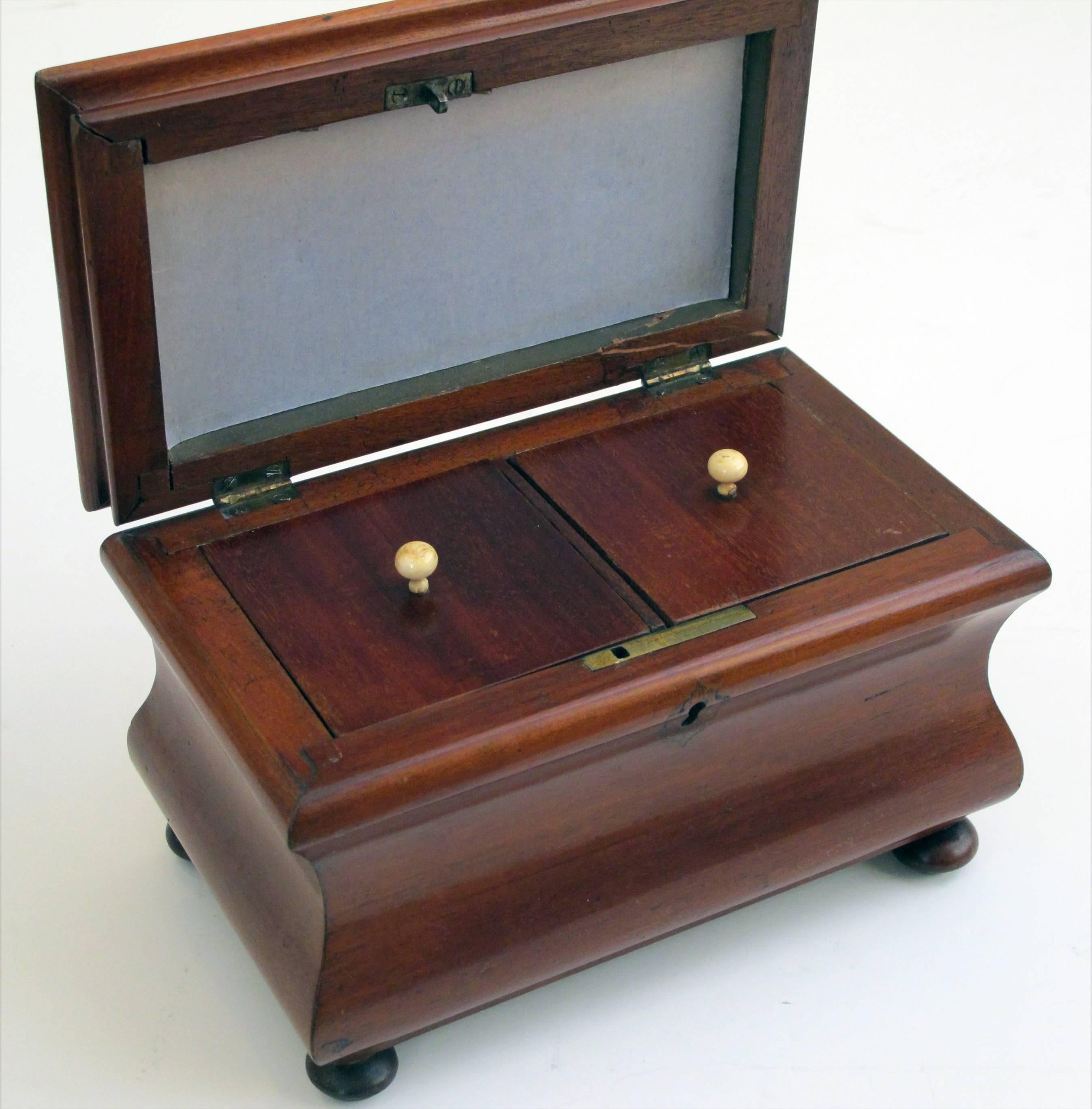A shapely English Victorian bombe-form mahogany tea caddy on bun feet; the shaped lid opening to reveal double compartments; resting on a waisted body all-over bun feet.