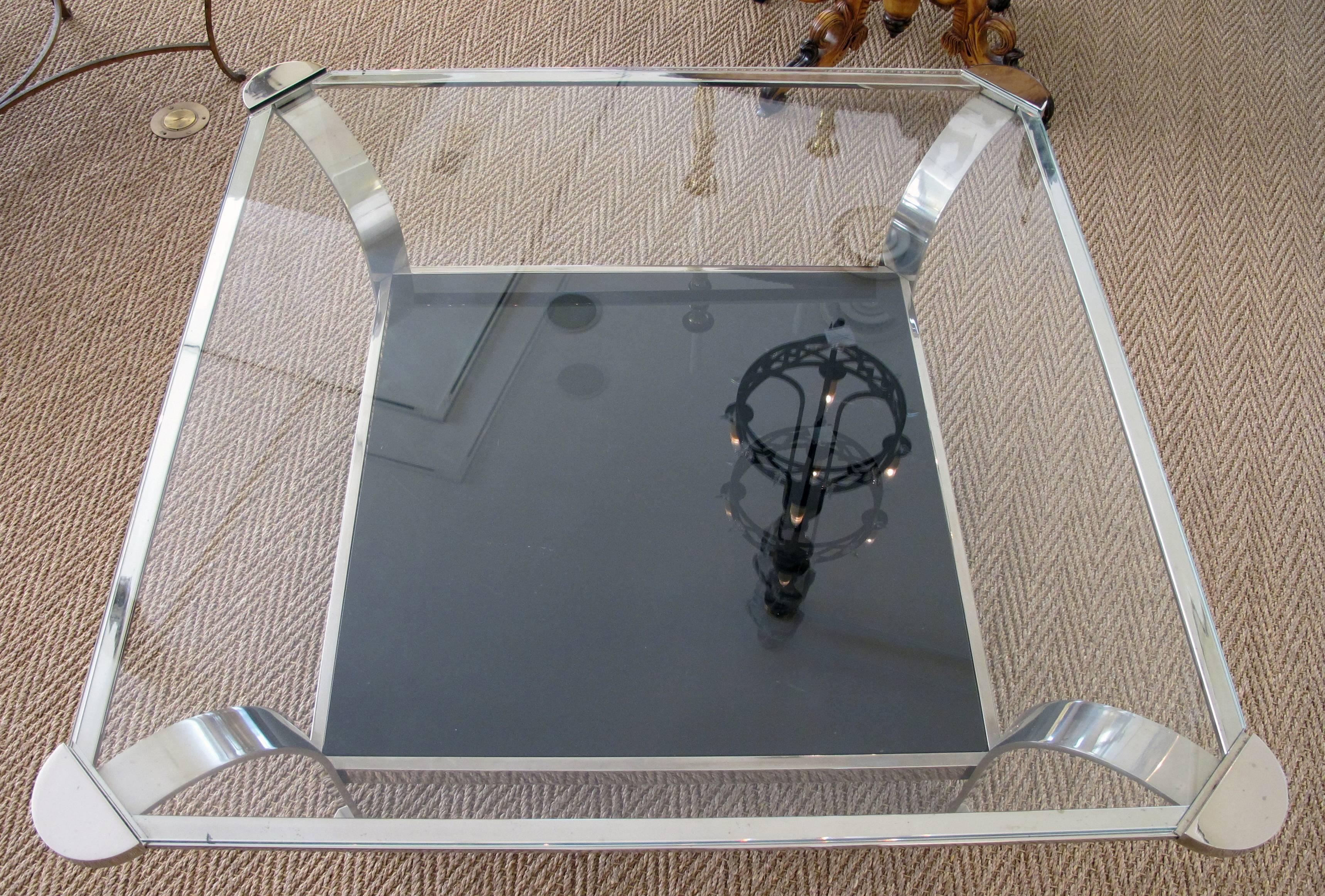 A large-scaled and shapely French 1970s steel square-form coffee table with clear glass top and black glass lower shelf; the square top with exaggerated rounded corners inset with a clear glass top; raised on incurved steel supports all joined by a