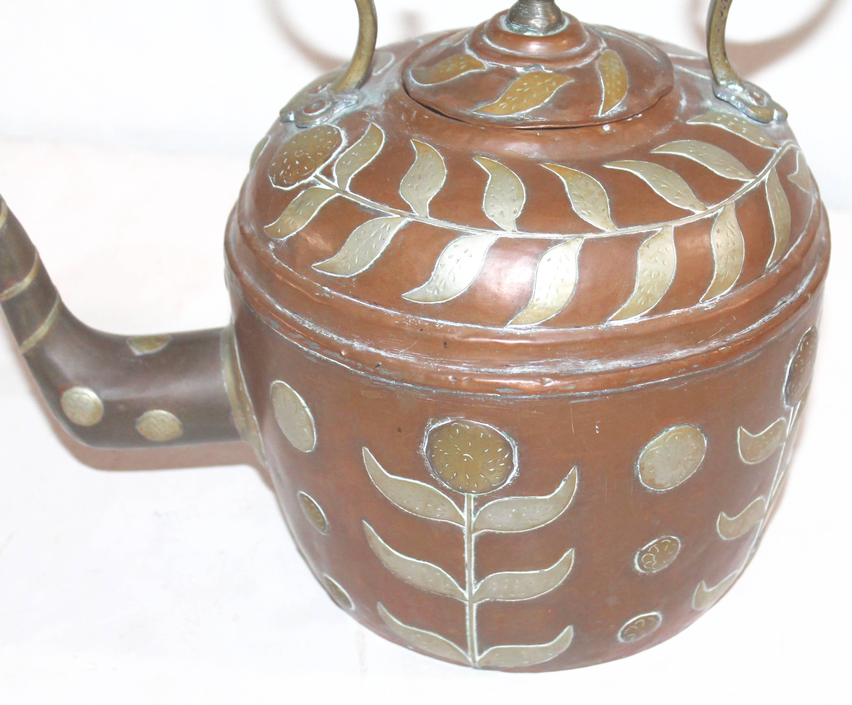 This highly decorated handmade copper kettle has appliqued sunflowers in silver and brass cutouts. The handle and stem is wrapped in silver applique work. The condition is very good.