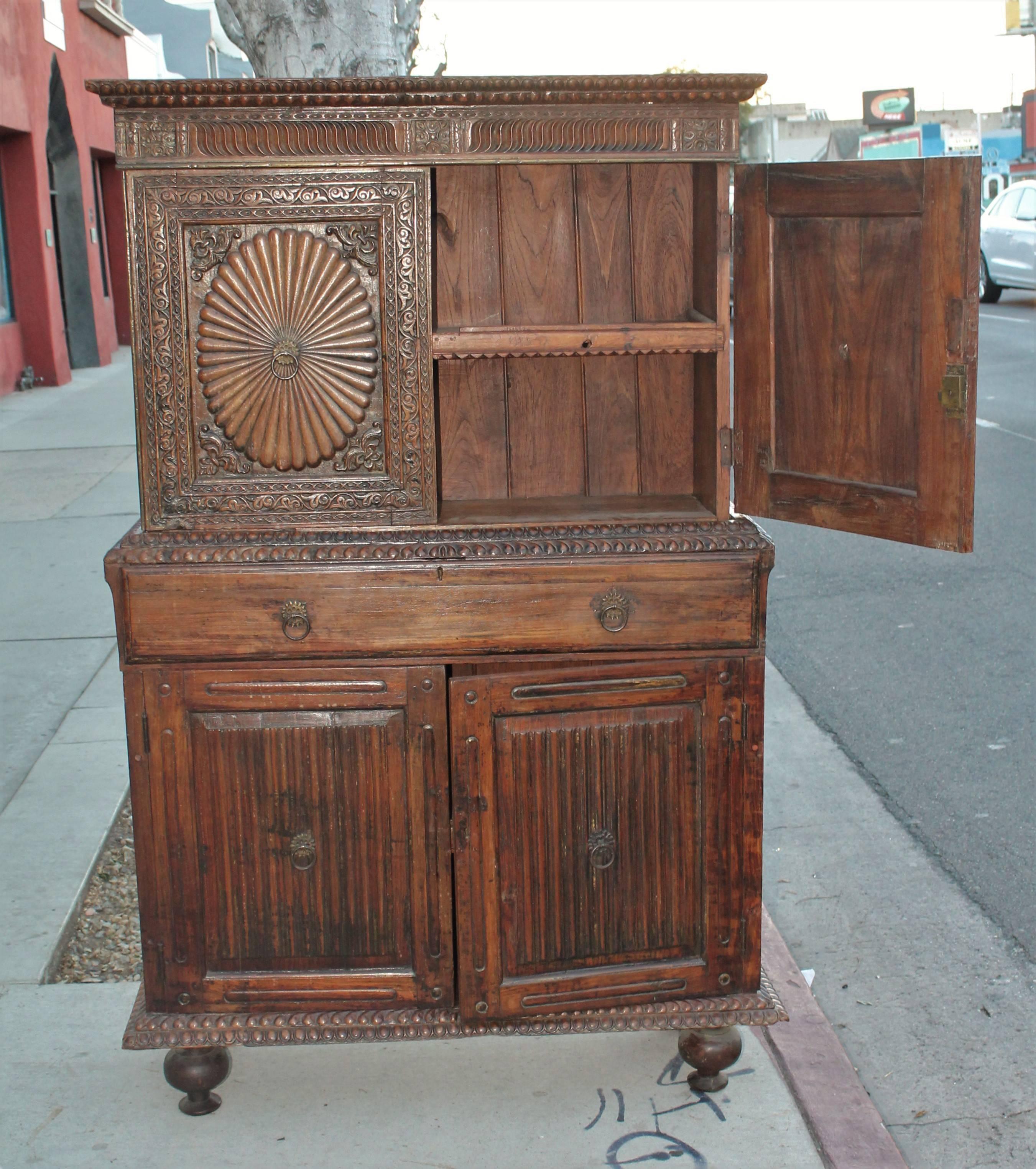 This amazing hand-carved teak wall cabinet is in two pieces and in very good condition. This cabinet is all hand-carved and early wood peg construction. The center drawer is a pulled down desk with small drawers inside. Minor wear consistent with