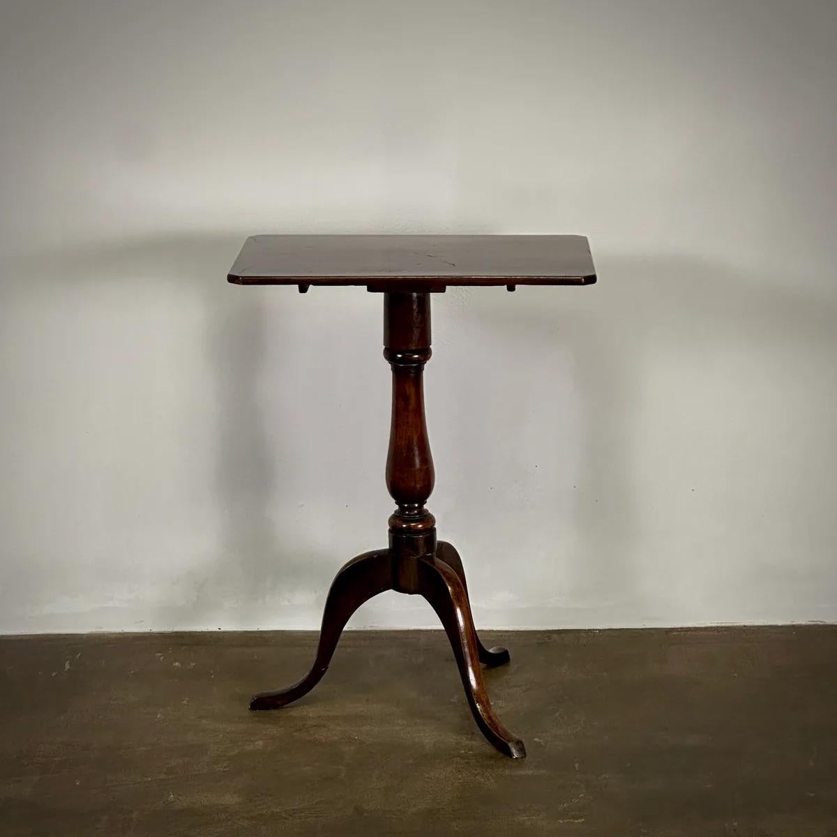 This Georgian tilt-top side table boasts a square mahogany surface and a graceful tripod base, typical of the refined styles prevalent in early 19th-century England. Its ability to tilt adds versatility while the rich patina reflects its storied