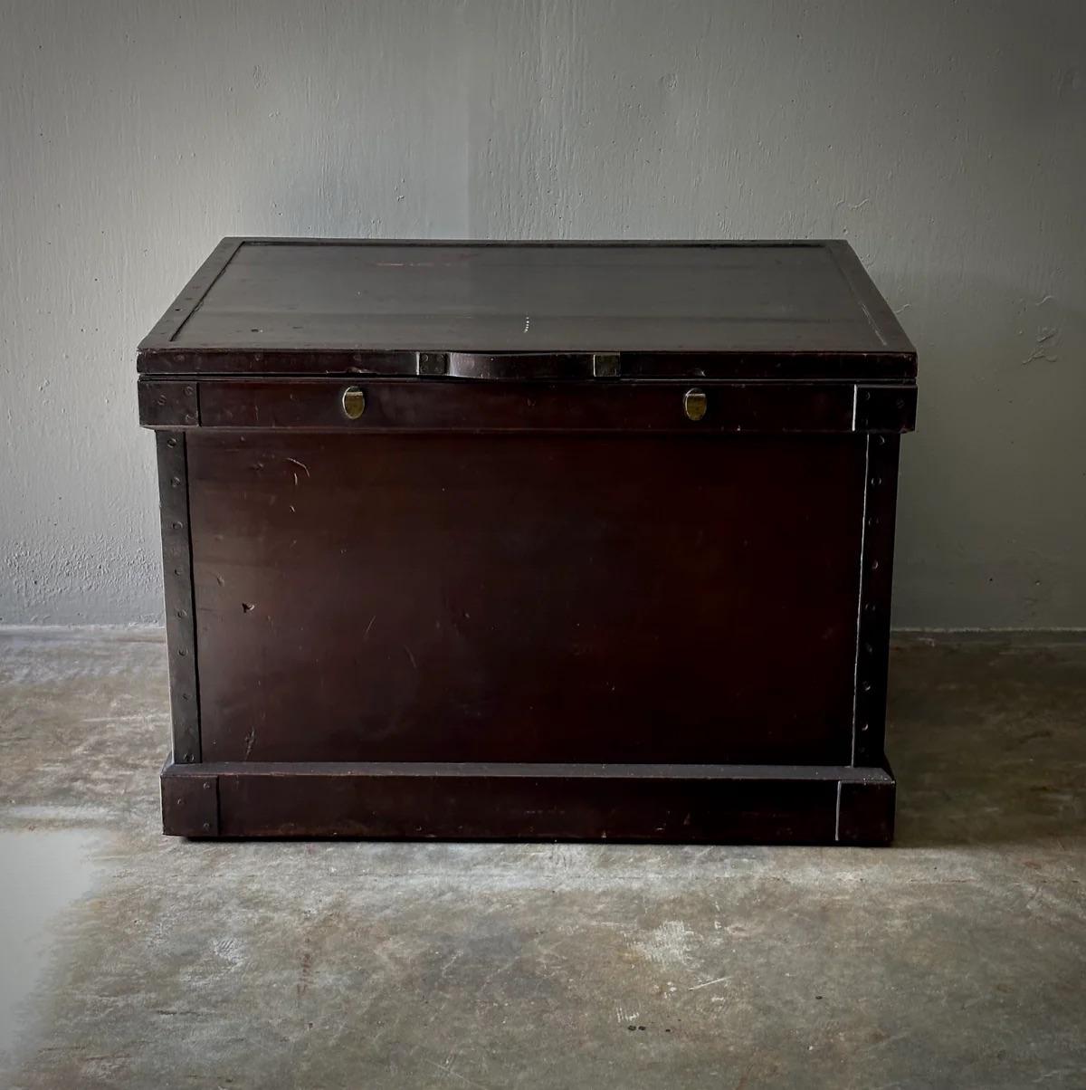 Questa grande cassapanca in legno di canfora incarna un fascino robusto, con la sua profonda tinta mogano e la sua struttura robusta. Presenta pratiche maniglie in pelle e un esterno in ferro borchiato che ne esaltano il fascino antico. Adatto per