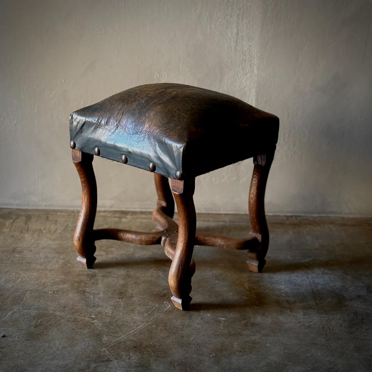 Tabouret sculptural en noyer aux courbes riches et à l'assise en cuir profondément patiné, fixé par une garniture de tête de clou proéminente. Les pieds et le croisillon, sculptés à la main, présentent une menuiserie experte et une asymétrie