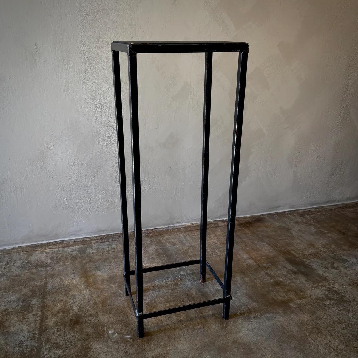 This industrial display pedestal or plinth is a minimalist yet utilitarian piece, designed to elevate sculptures, artifacts, or decorative objects with an understated elegance. Its slim metal frame and matte black surface create a modern aesthetic