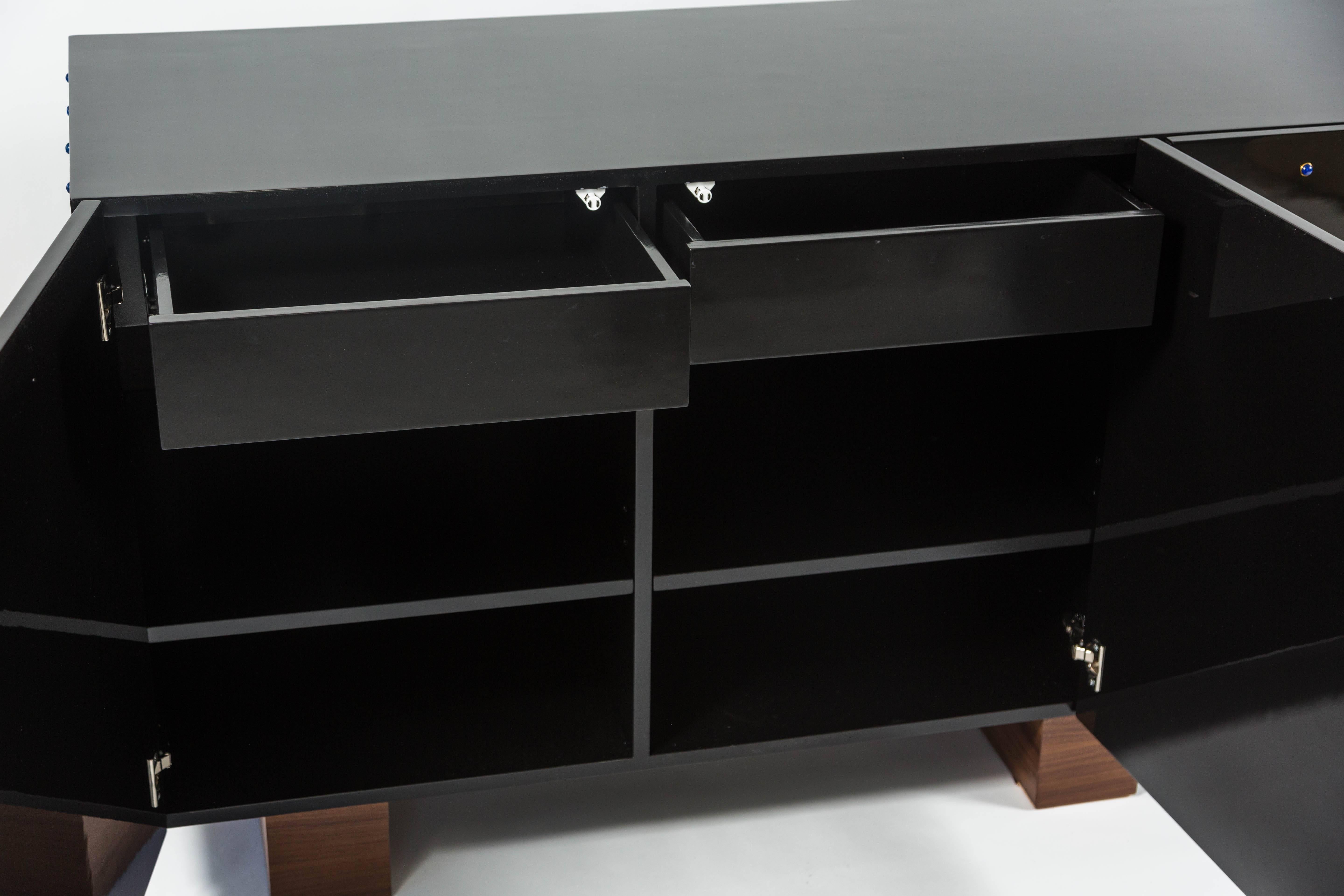 Un'elegante credenza a quattro ante in lacca nera, tempestata di cabochon di lapis, che poggia su tre guide in noce. Il mobile è inoltre dotato di una ferramenta con chiusura a sfioramento e di due cassetti all'interno. Questo design può essere