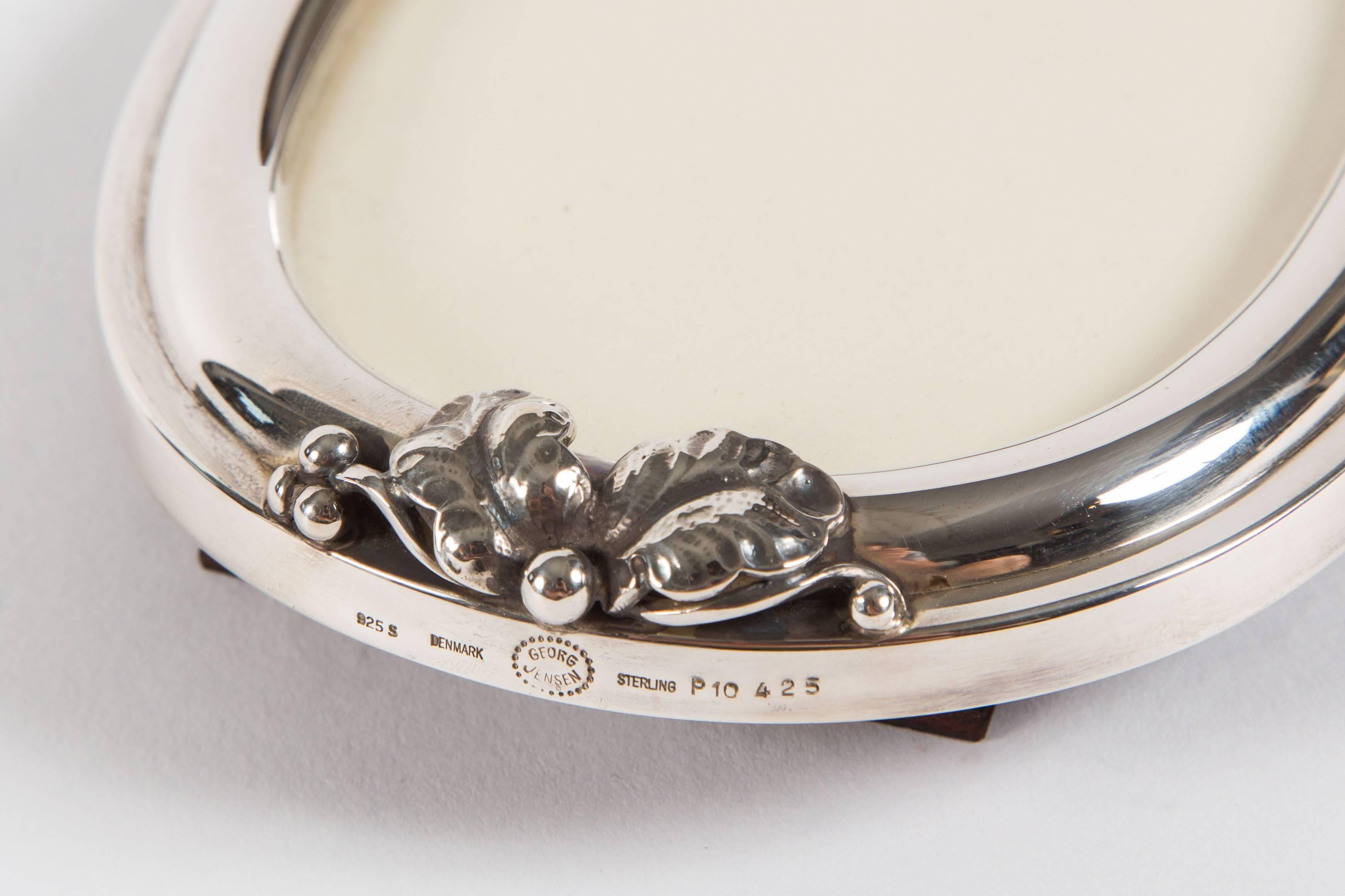 Delightful frame by renowned Danish manufacturer Georg Jensen. Sterling silver. With floral ornamentation at the base of the frame. Bears Georg Jensen, and additional hallmarks beneath the ornamentation, on the edge of the frame. Swedish date mark