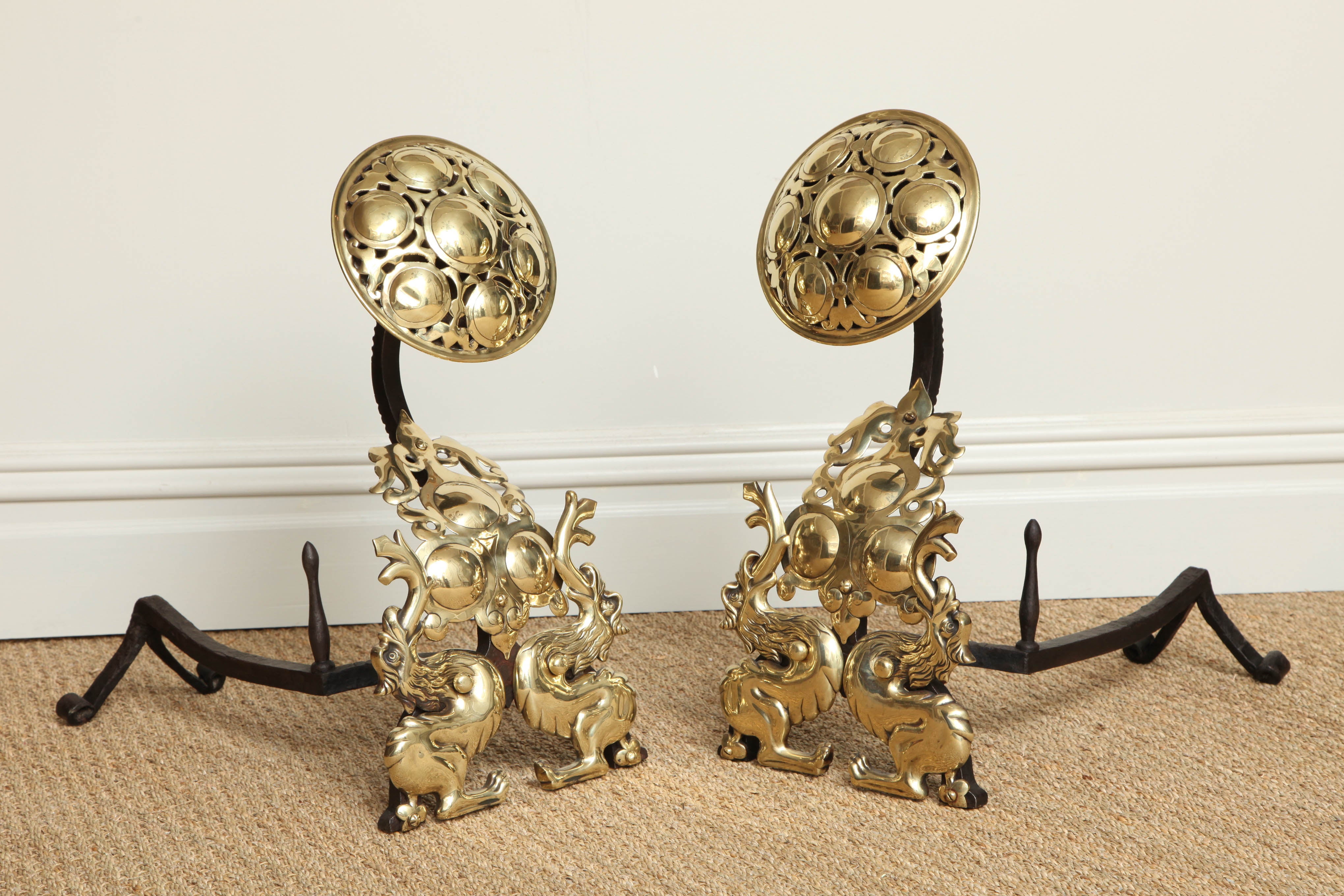 Striking and distinctive pair of English Arts and Crafts andirons, the pierced brass tops with embossed cabouchons over swan necks flanked by fire breathing griffins or wyverns bestride roundel embellished cartouches, with nicely worked wrought iron
