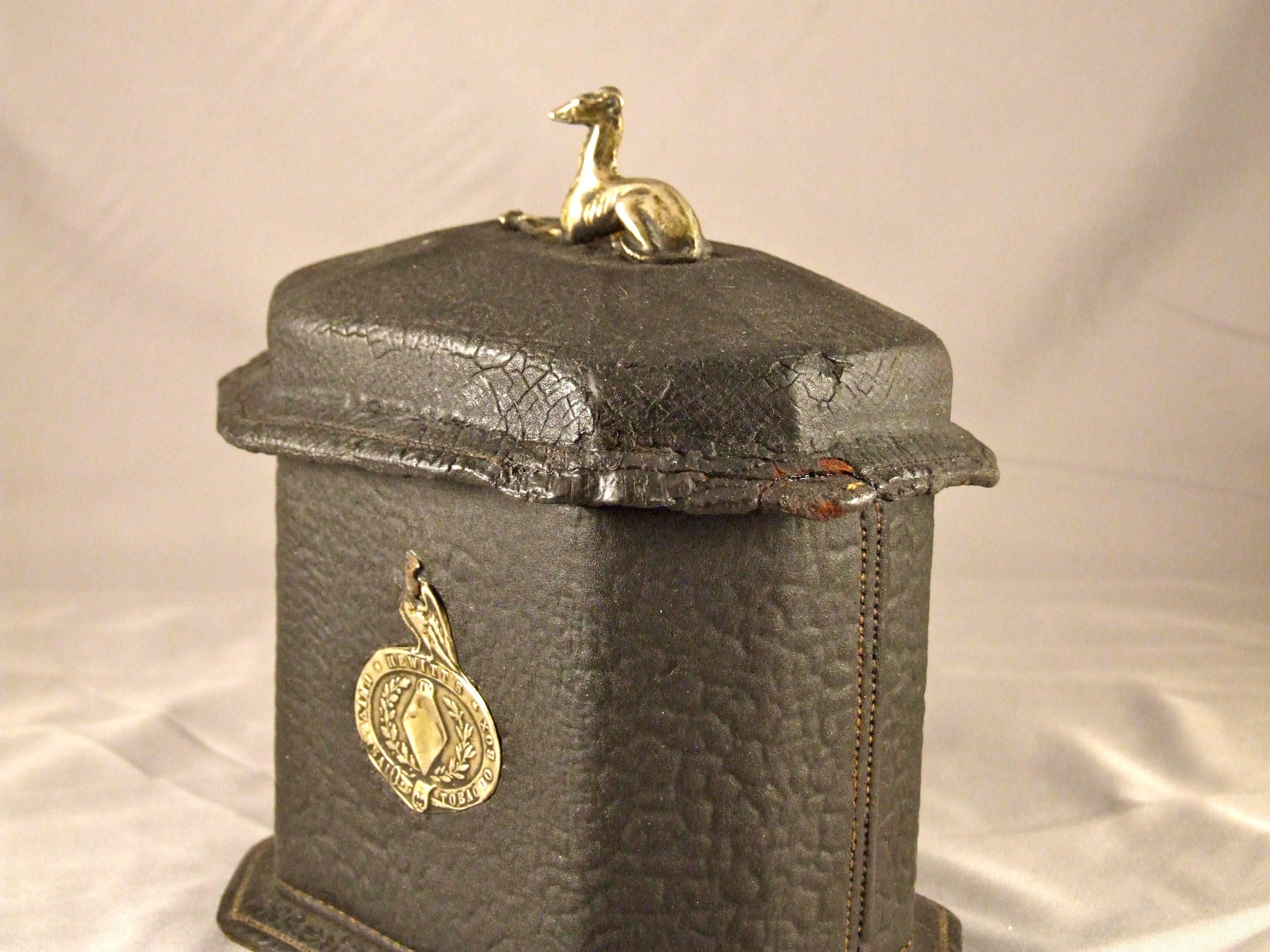 An English 19th century tobacco caddy of tin covered in stitched shagreen, of oblong hexagonal form with a lid surmounted by a silver whippet, retaining remnants of label 