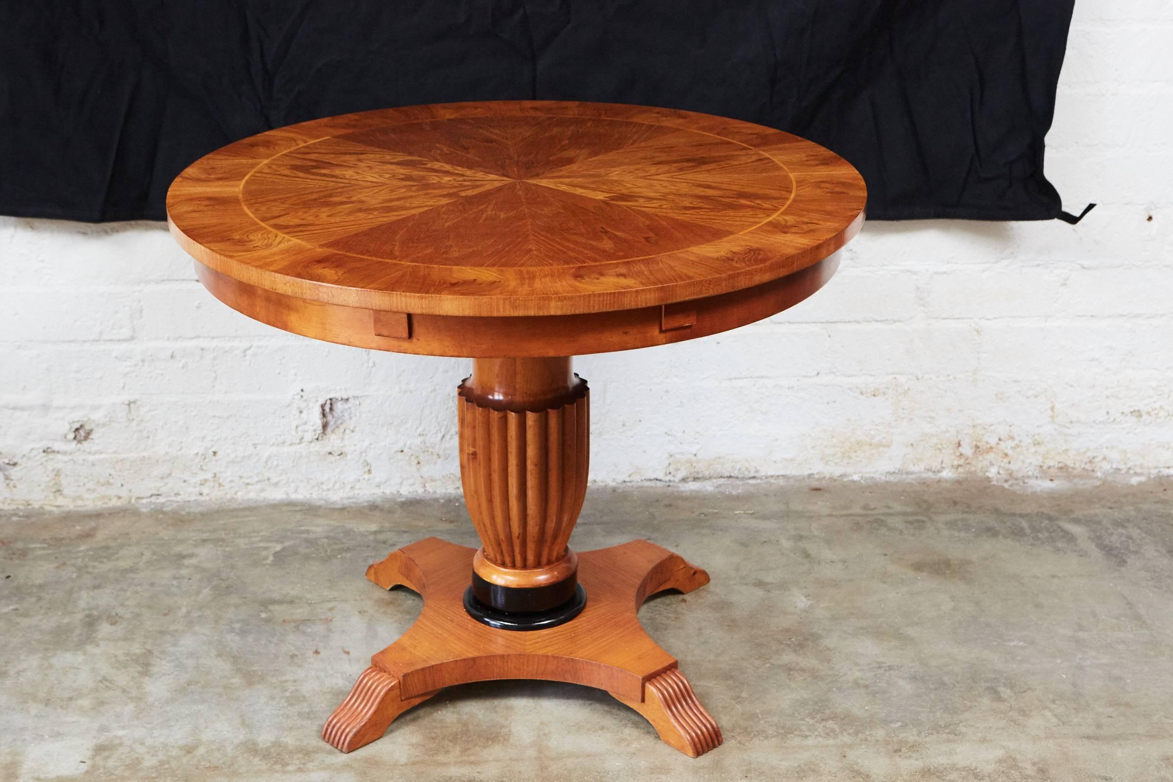 This handsome Biedermeier pedestal table has a round veneered top with circular banding.The top is supported by a fluted central stem with a black painted detail on a platform base with four scrolled feet.

