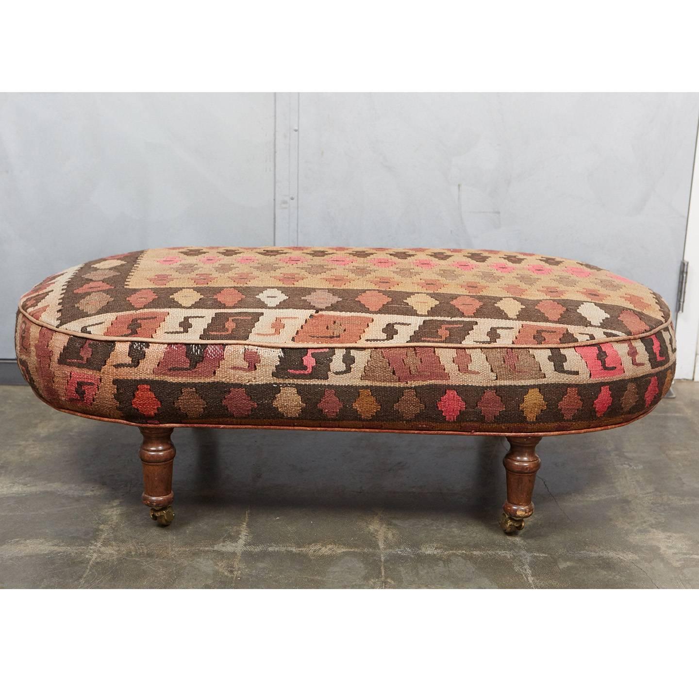 This interesting oval ottoman was custom-made in England in the 1980s. It has turned wood feet on casters and wheels. It is upholstered with a beautifully woven 1920s Kilim rug and trimmed with distressed leather piping. See photos for details of