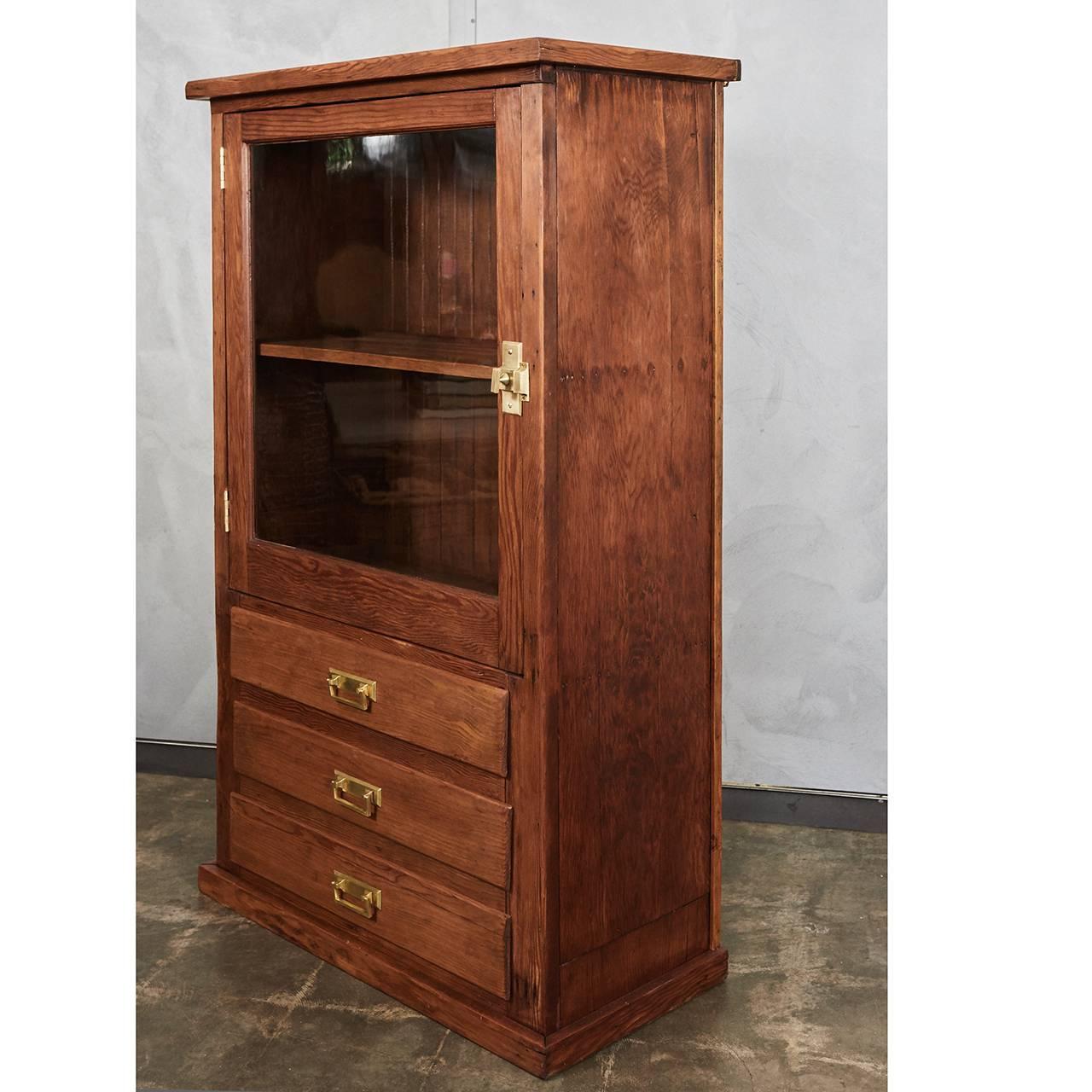 This handsome pine cabinet from the early 20th century is well suited for a variety of settings. The piece has the original brass fixtures that have been polished to highlight their appearance. The body has a shelf and a glass door above three