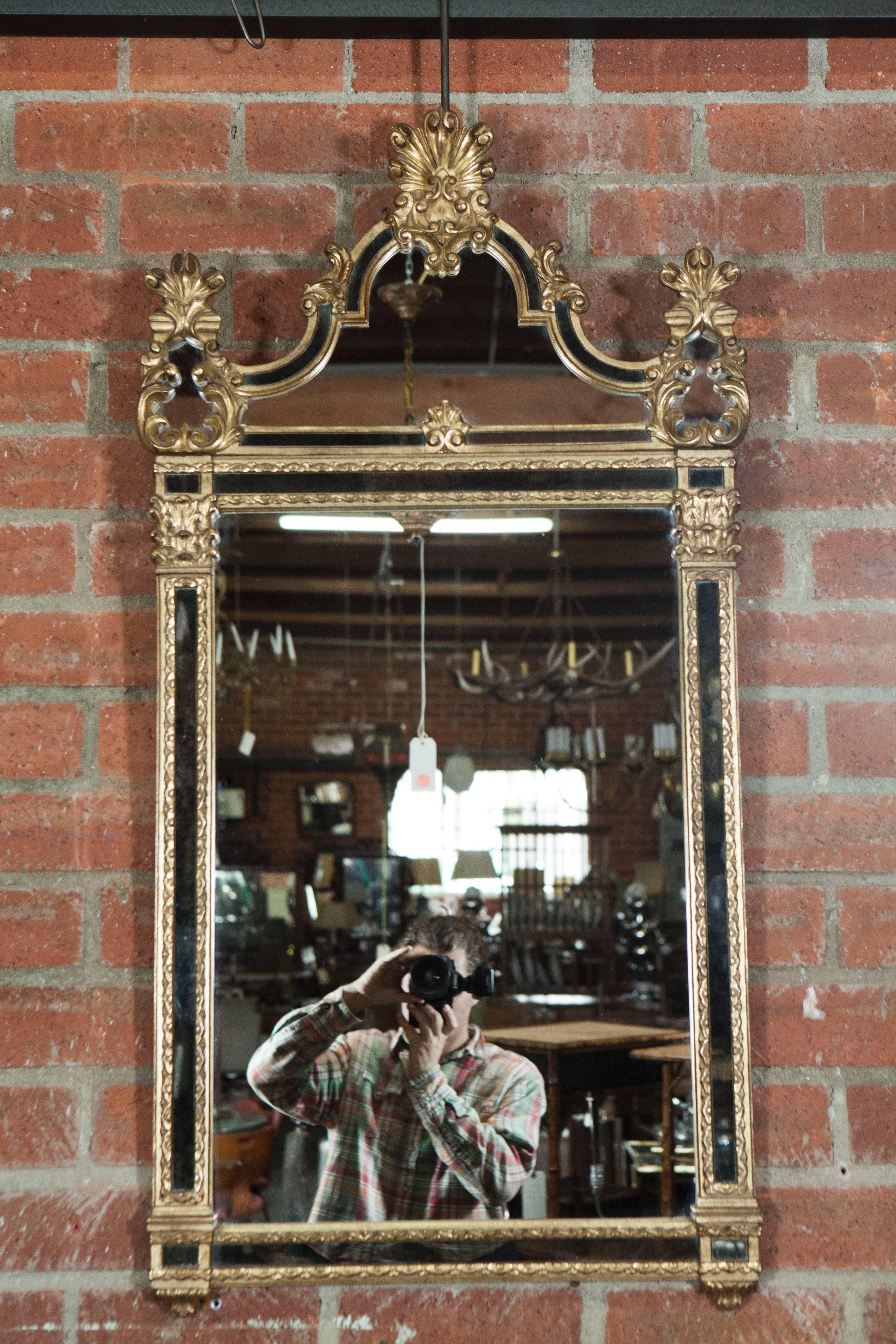 A beautifully hand-carved and gilt finished French Regency style framed mirror from the early 20th century. The mirror has a dome shaped capital decorated with with multiple c carved designs, set above a square frame-within-frame border of flora and