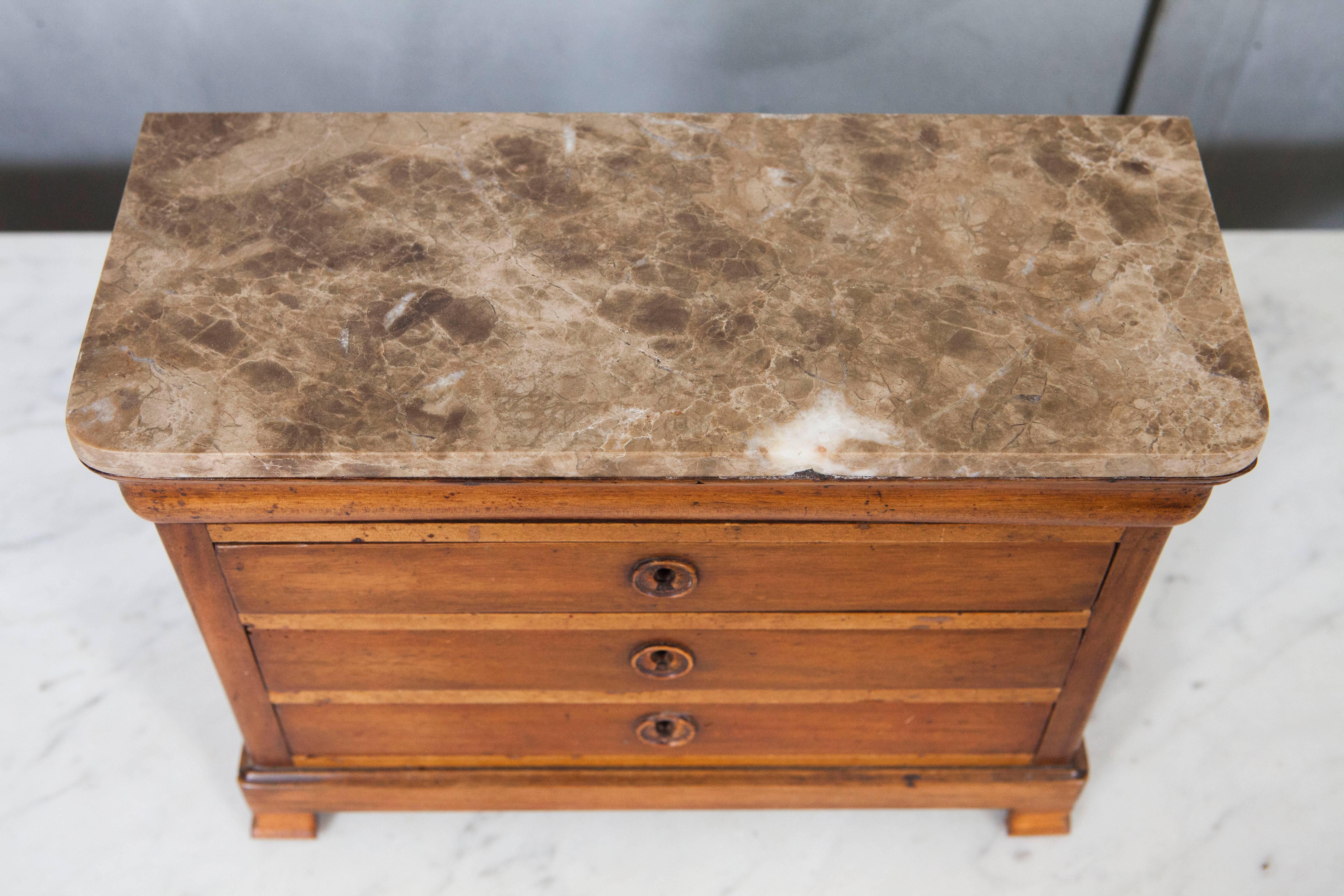 This handsome miniature chest of drawers has a marble top, pedestal feet and wooden escutcheons. The piece has dovetail construction and brass locks.