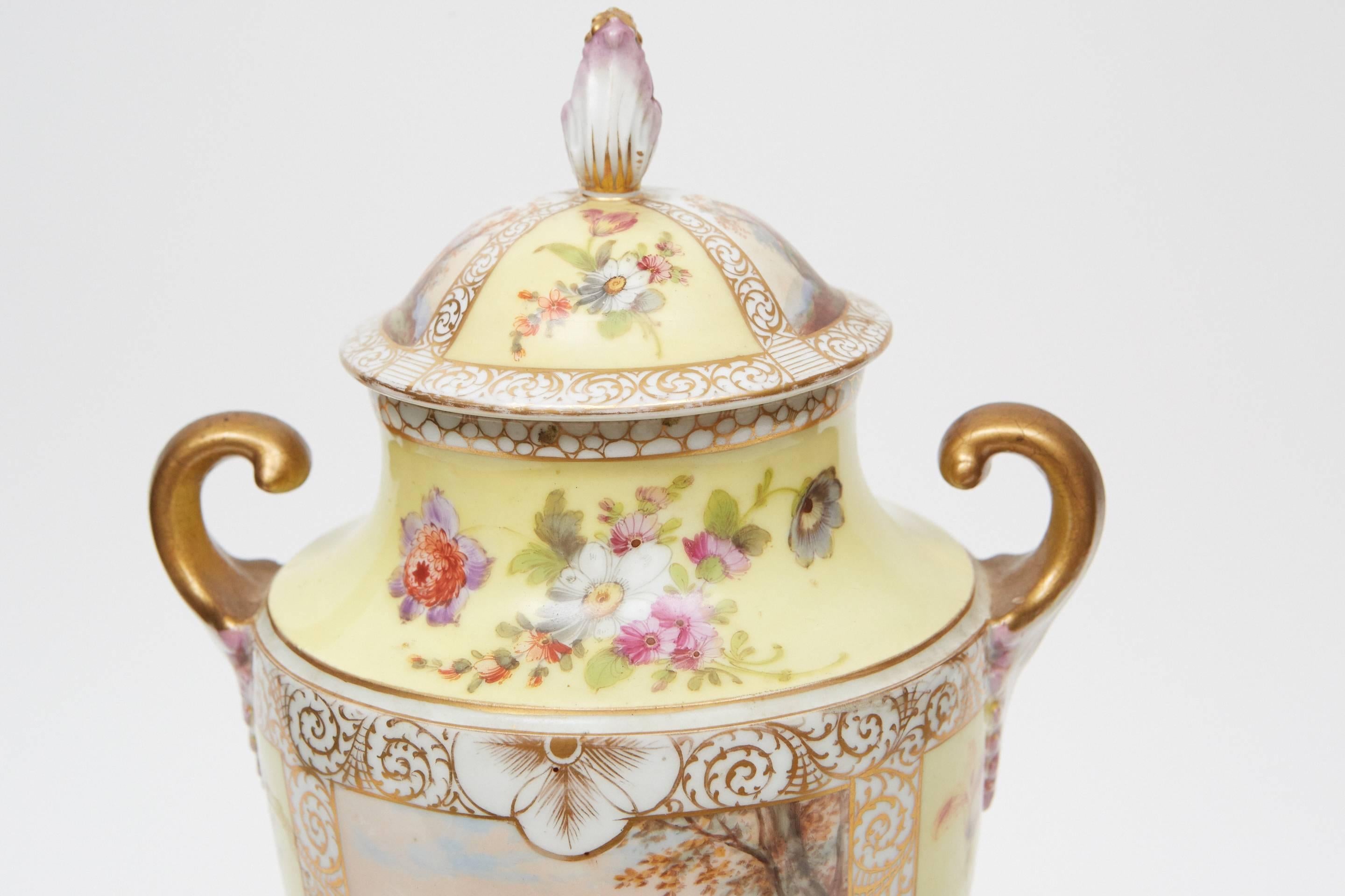 This Dresden hand-painted porcelain urn on plinth has floral and figurative elements throughout with two large scenes of couples on each side. The piece has gold painted decorative boarders and and two gold and rose hand-painted handles. The maker's