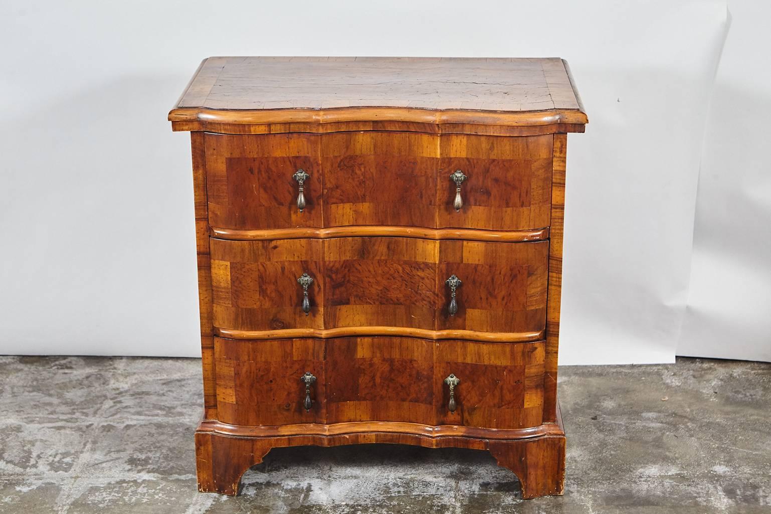 This pair of beautifully veneered serpentine front nightstands have three drawers and brass drop handles. The nightstands have paneled sides, wide banding on the surface top and sit on four shaped feet paneled sides.