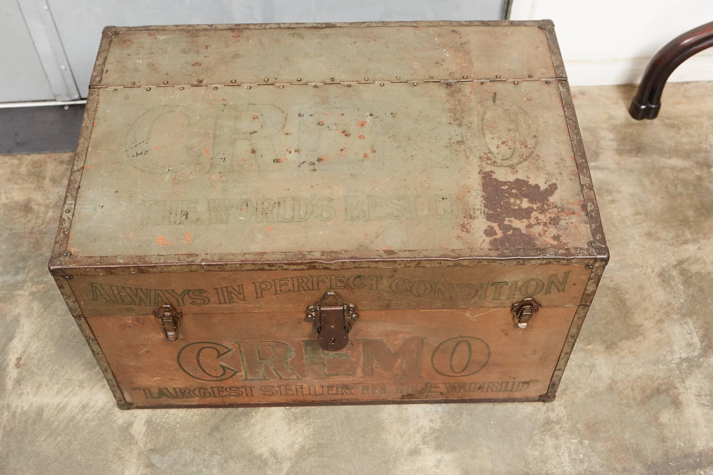 This metal Cremo cigar trunk has a beautiful patina with worn orange, green and black coloring and lettering. The trunk was used to store and ship cigar boxes as indicated by the vented portal on the side. The trunk has several slogans and logos