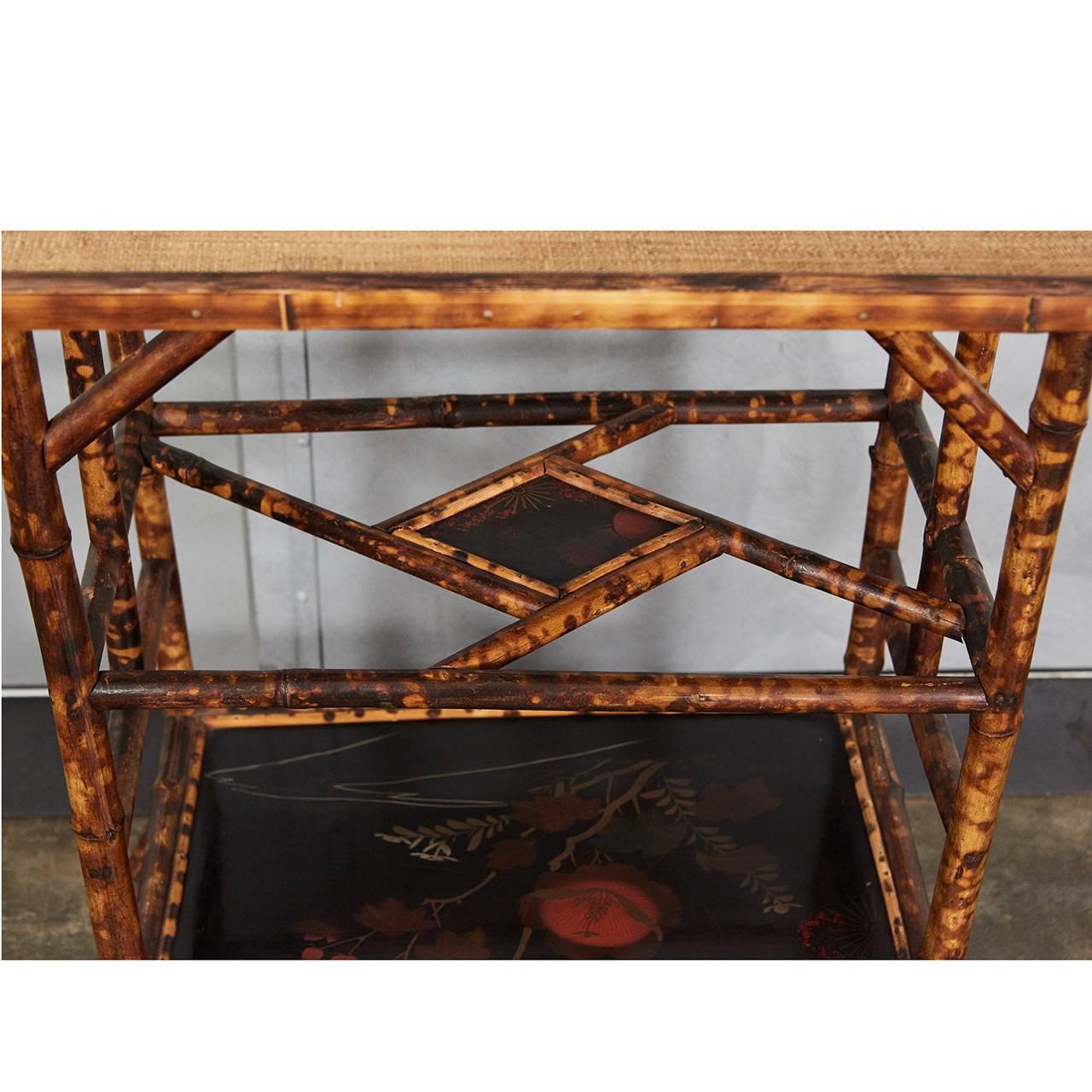 This exquisite English Victorian tiger bamboo table has intricate two lacquered panels and intricate lattice supports. Between the top and the lower shelf are a set of supports holding an inset diamond shaped lacquered panel. The lower shelf has a