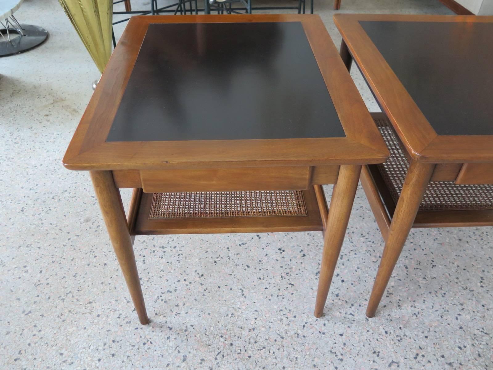 A pair of American of Martinsville occasional tables. Pull out drawer, caned shelf underneath. Walnut/black micarta tops.