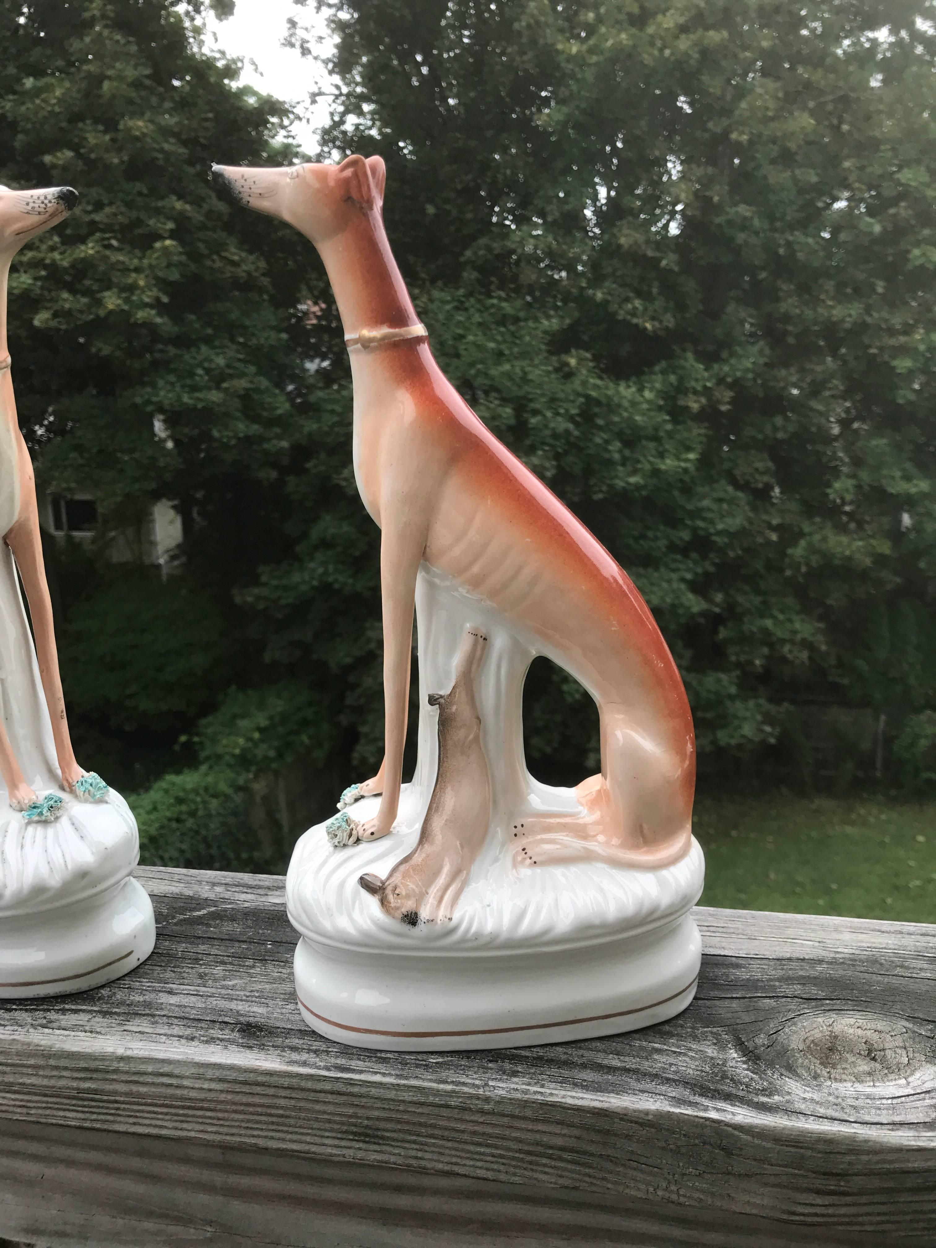 Pair of English Staffordshire porcelain greyhounds or whippets shown seated in stately fashion with the recently captured rabbit hanging from a tree stump. Each with a little tuft of grass in front of their paws. The utter charm of these figures is